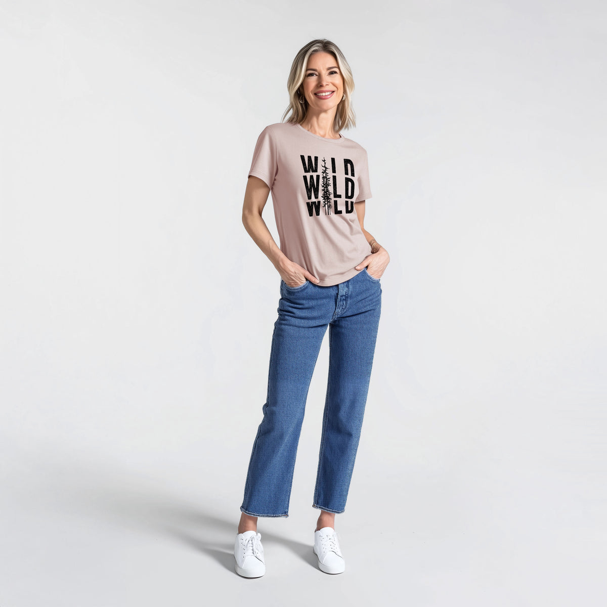 Woman wearing a pink gravel women's relaxed fit cotton t-shirt printed with bold black text reading WILD stacked three times, with a tall pine tree forming the letter I.