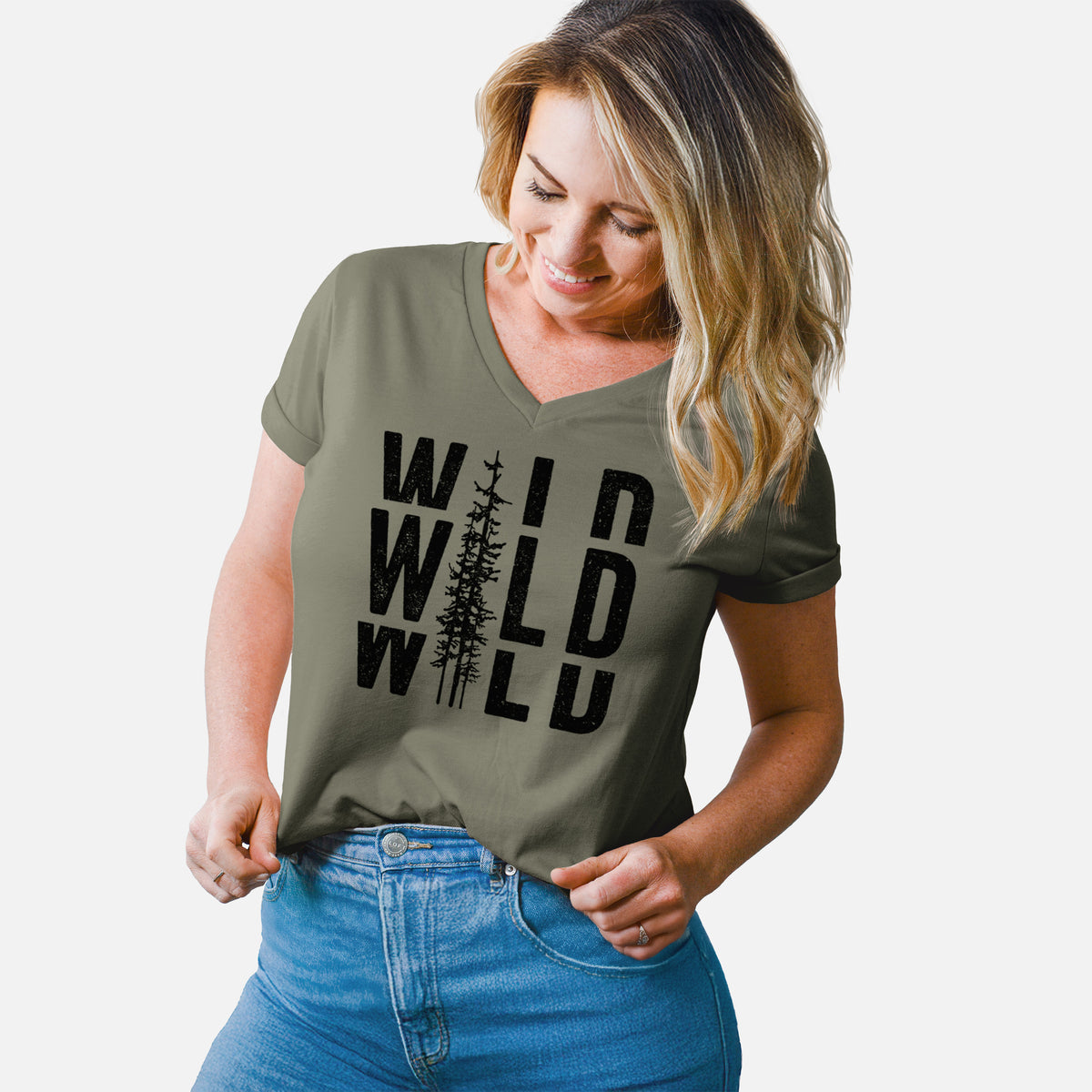 Woman wearing a military green women's relaxed fit cotton V-neck t-shirt printed with bold black text reading WILD stacked three times, with a tall pine tree forming the letter I.