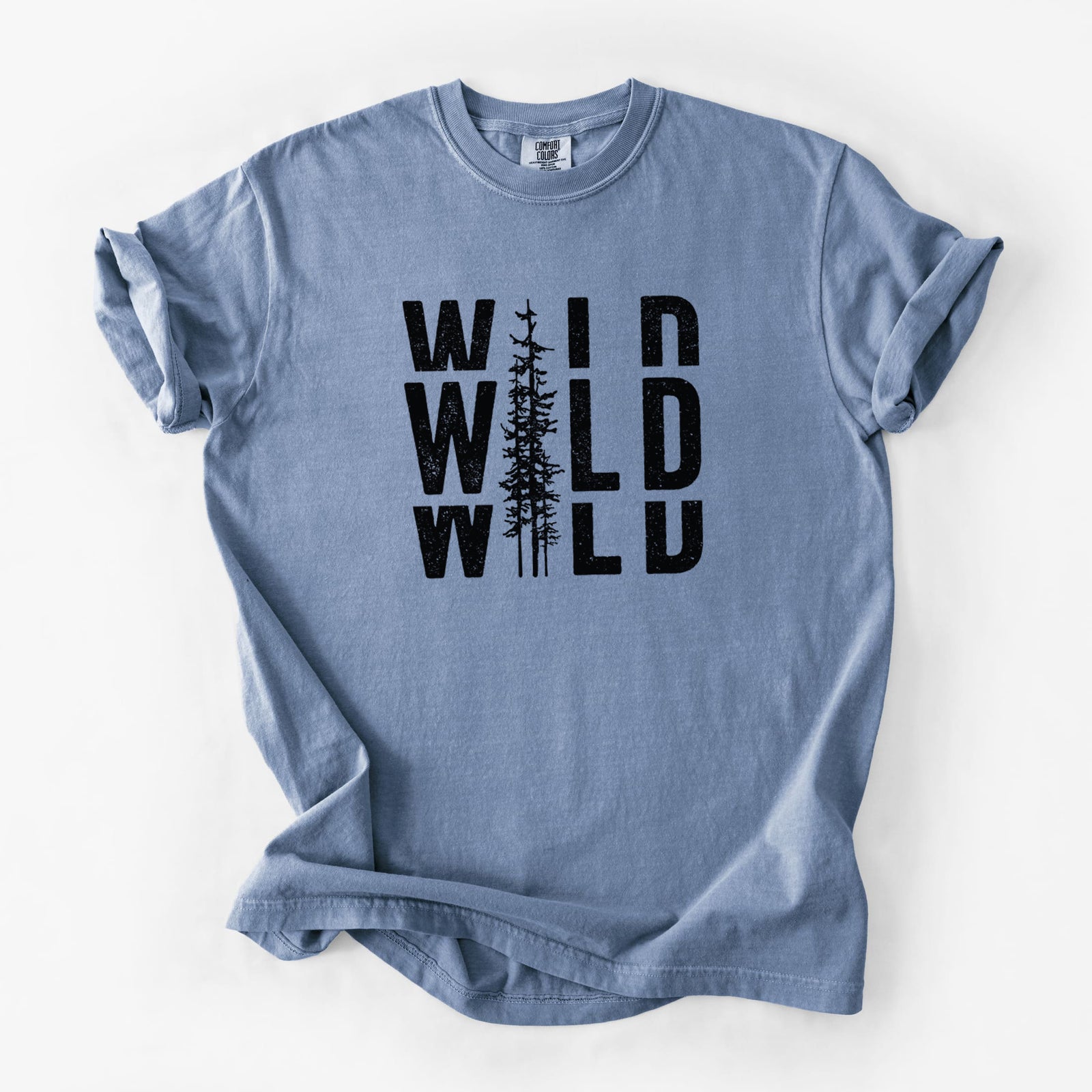 Blue jean Comfort Colors t-shirt laid flat, printed with bold black text reading WILD stacked three times, with a tall pine tree forming the letter I.