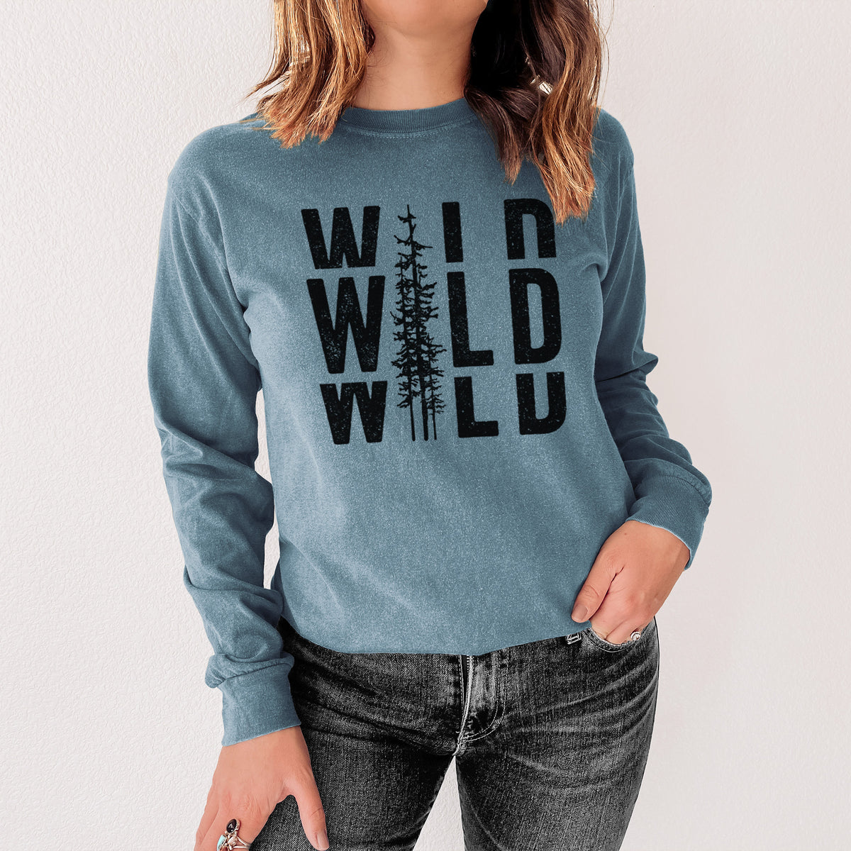 Steel blue Comfort Colors long sleeve shirt printed with bold black text reading WILD stacked three times, with a tall pine tree forming the letter I, shown in a lifestyle photo.