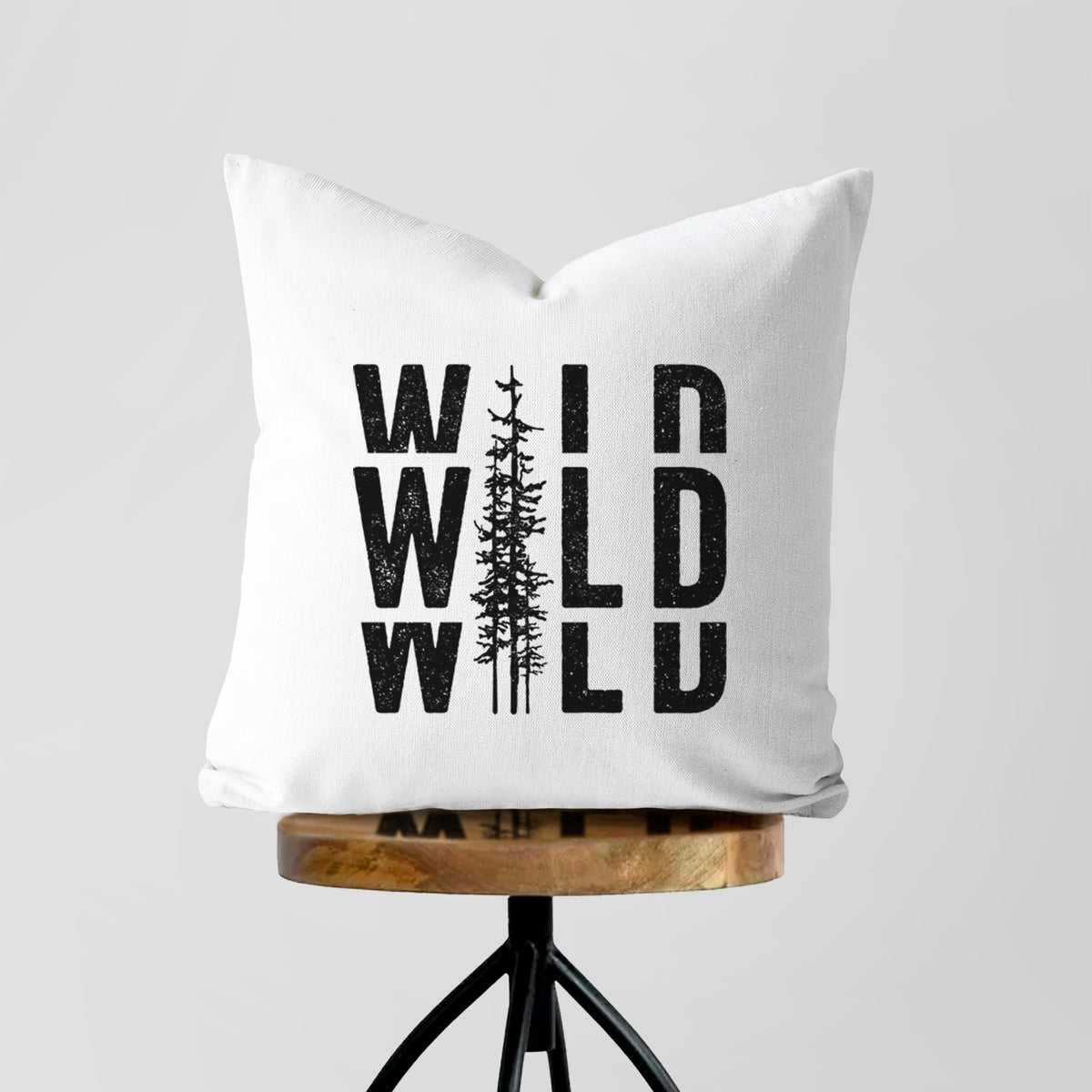 White pillow cover printed with bold black text reading WILD stacked three times, with a tall pine tree forming the letter I, on a wooden stool.