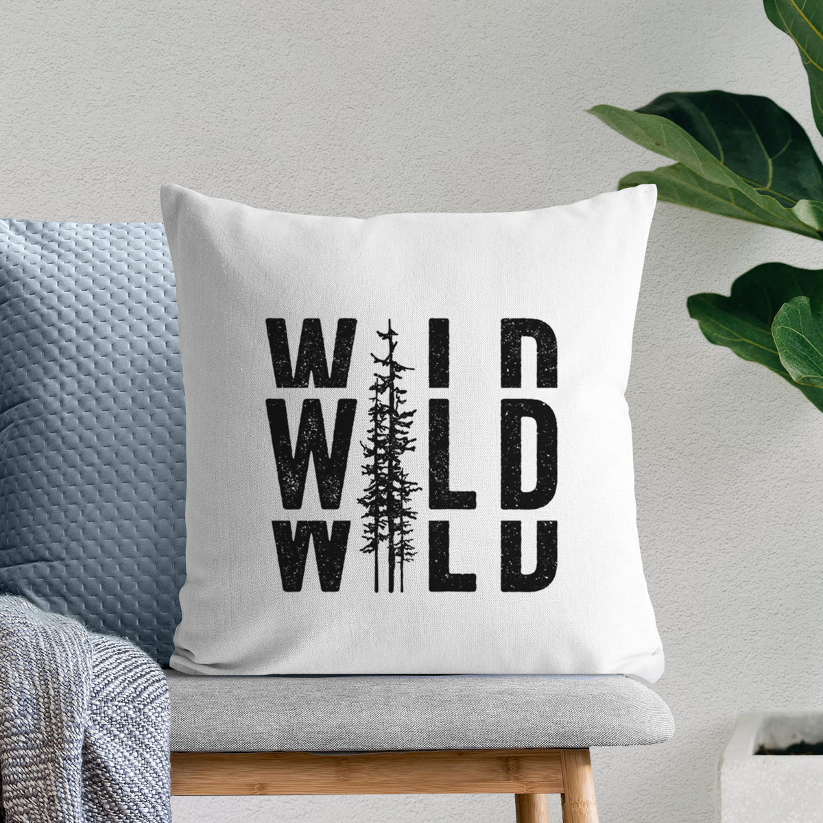 White pillow cover printed with bold black text reading WILD stacked three times, with a tall pine tree forming the letter I, on a bench beside plants.