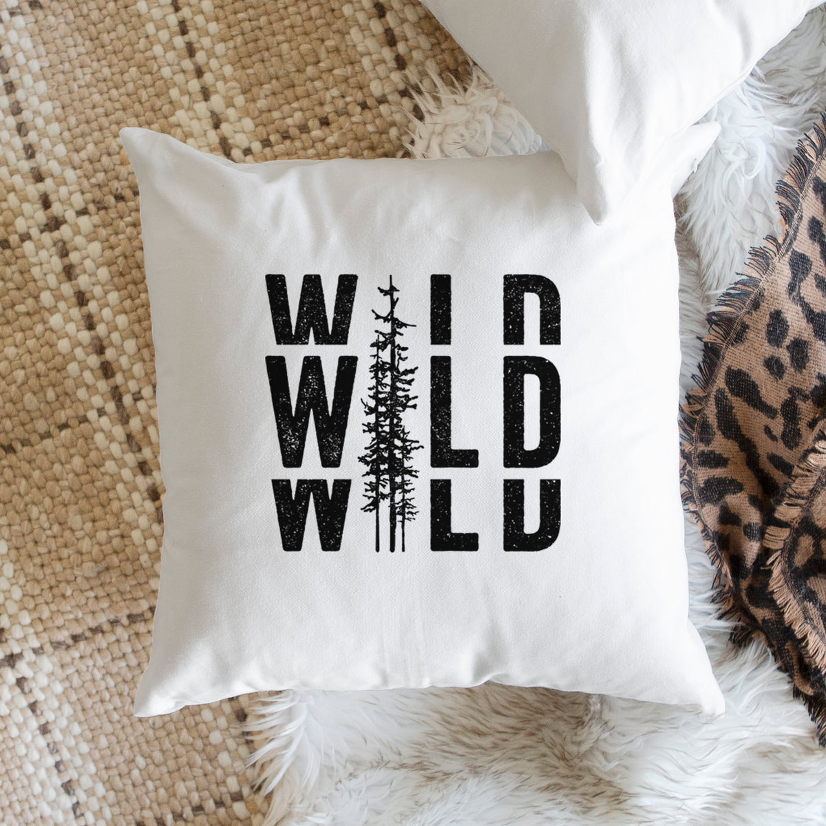 White pillow cover printed with bold black text reading WILD stacked three times, with a tall pine tree forming the letter I, on a woven chair.