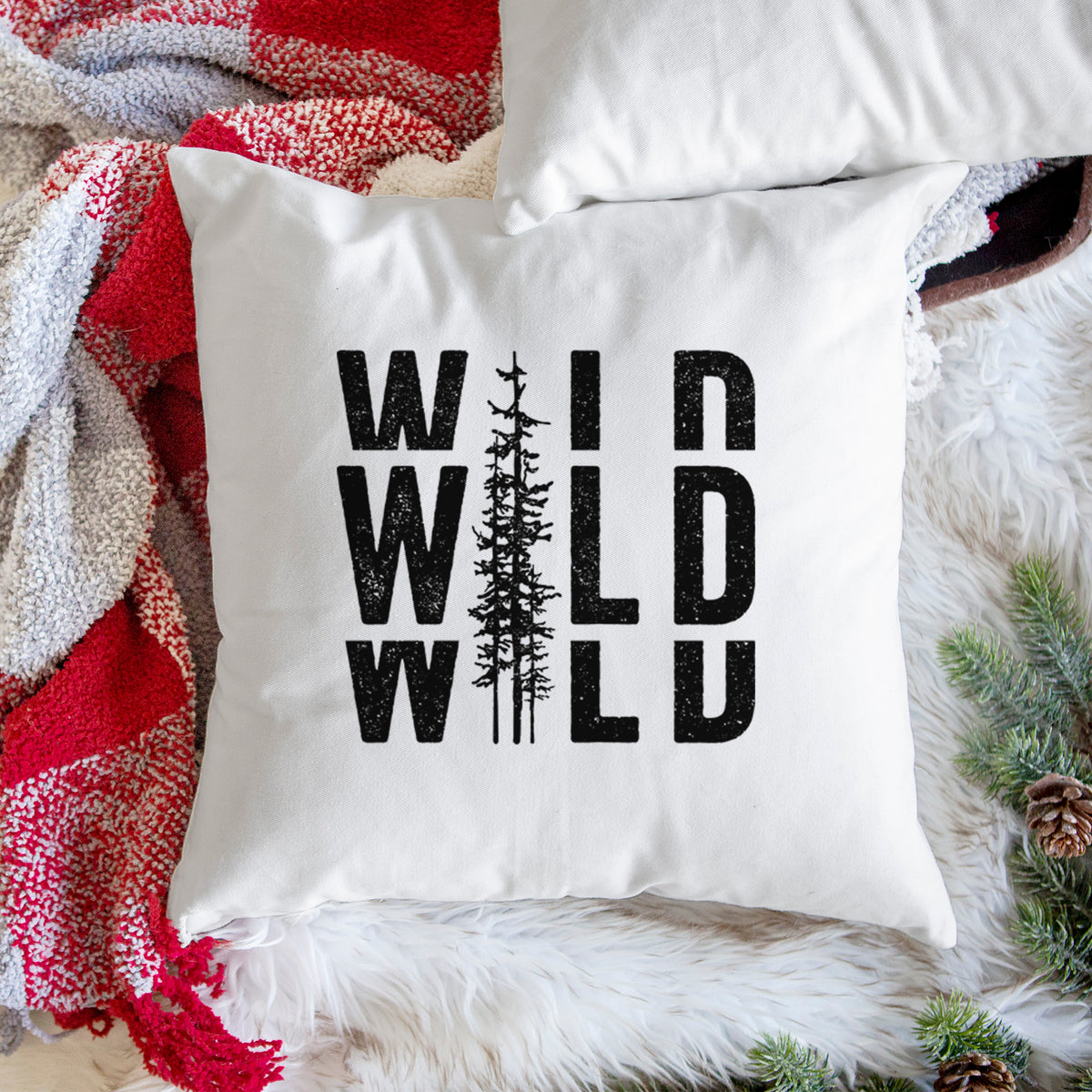 White pillow cover printed with bold black text reading WILD stacked three times, with a tall pine tree forming the letter I, on a woven chair.