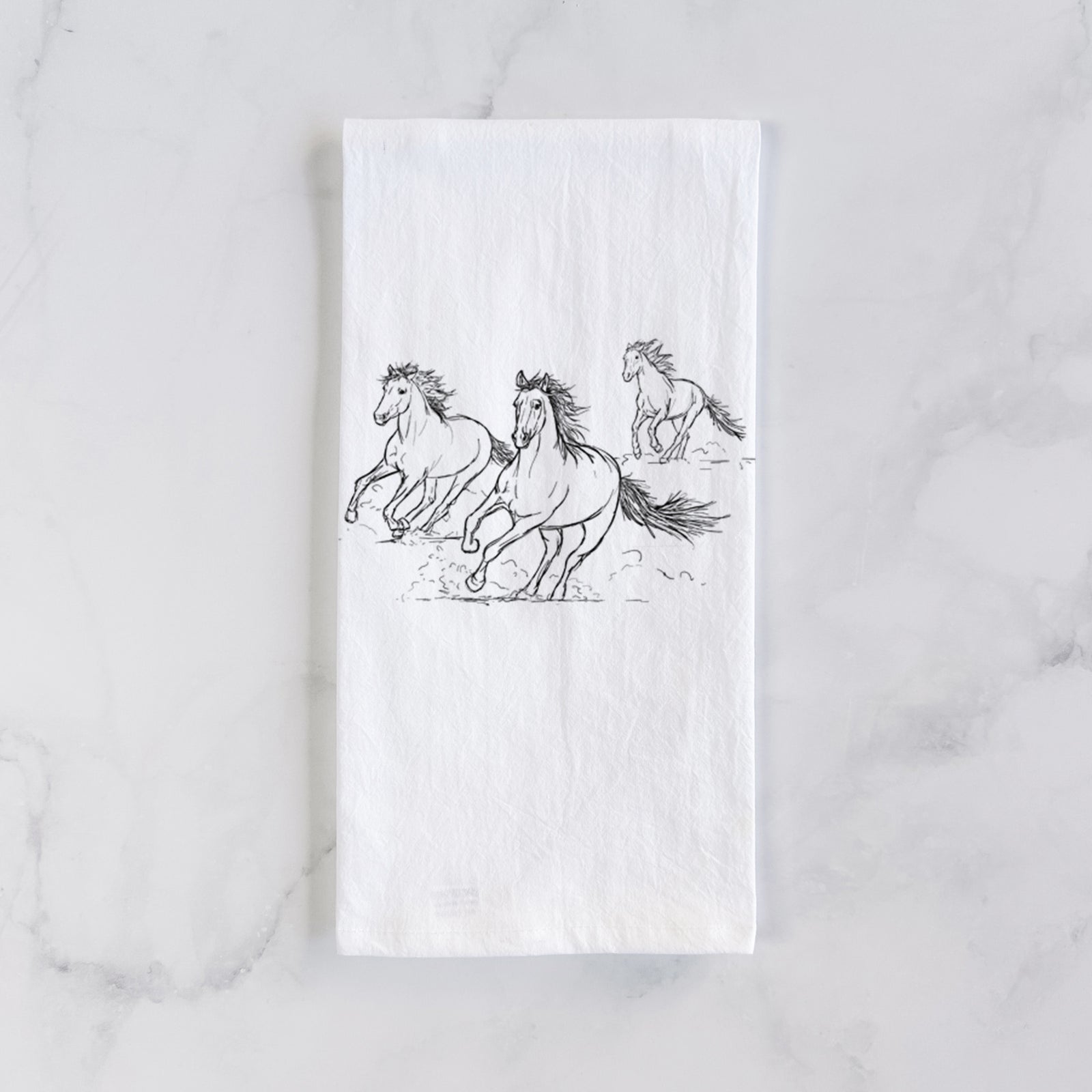 White cotton tea towel printed with a black ink illustration of three wild horses galloping, on a marble counter beside a round wooden cutting board and greenery.