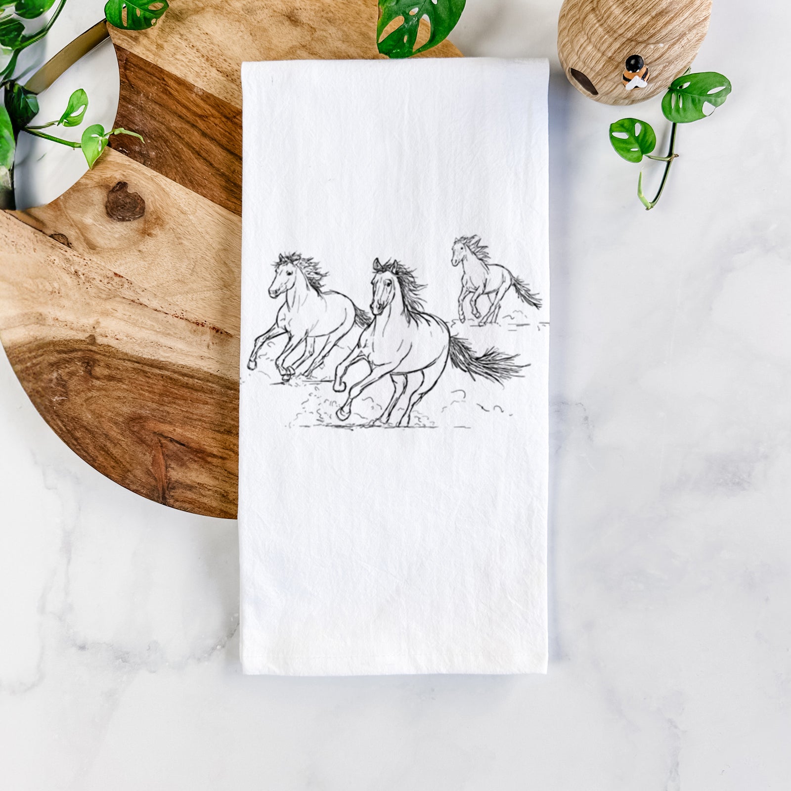 White cotton tea towel printed with a black ink illustration of three wild horses galloping, on a marble counter beside a round wooden cutting board and greenery.
