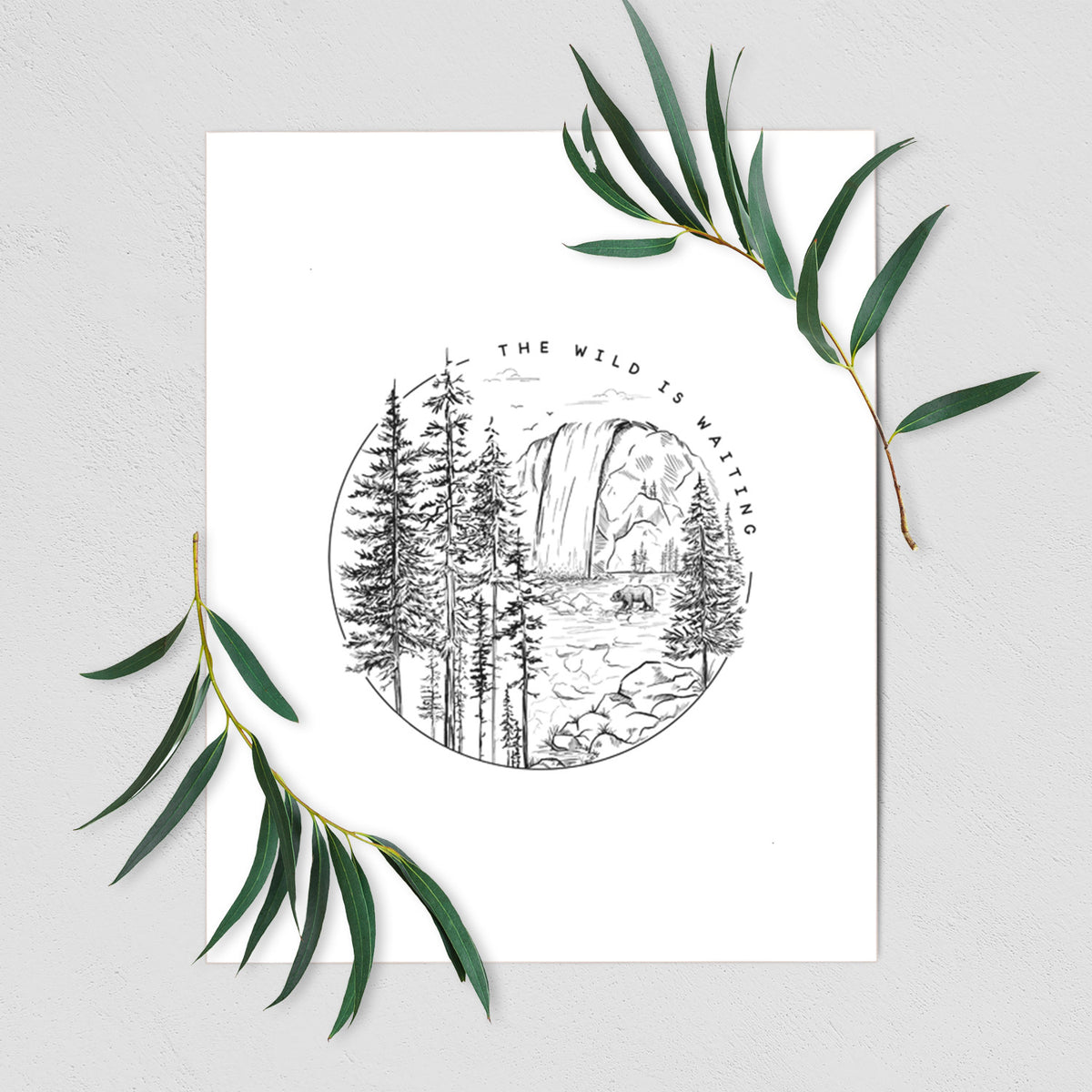 Art print of a round black ink scene of a waterfall, pine trees and a river, with the words The Wild Is Waiting, shown with eucalyptus leaves.