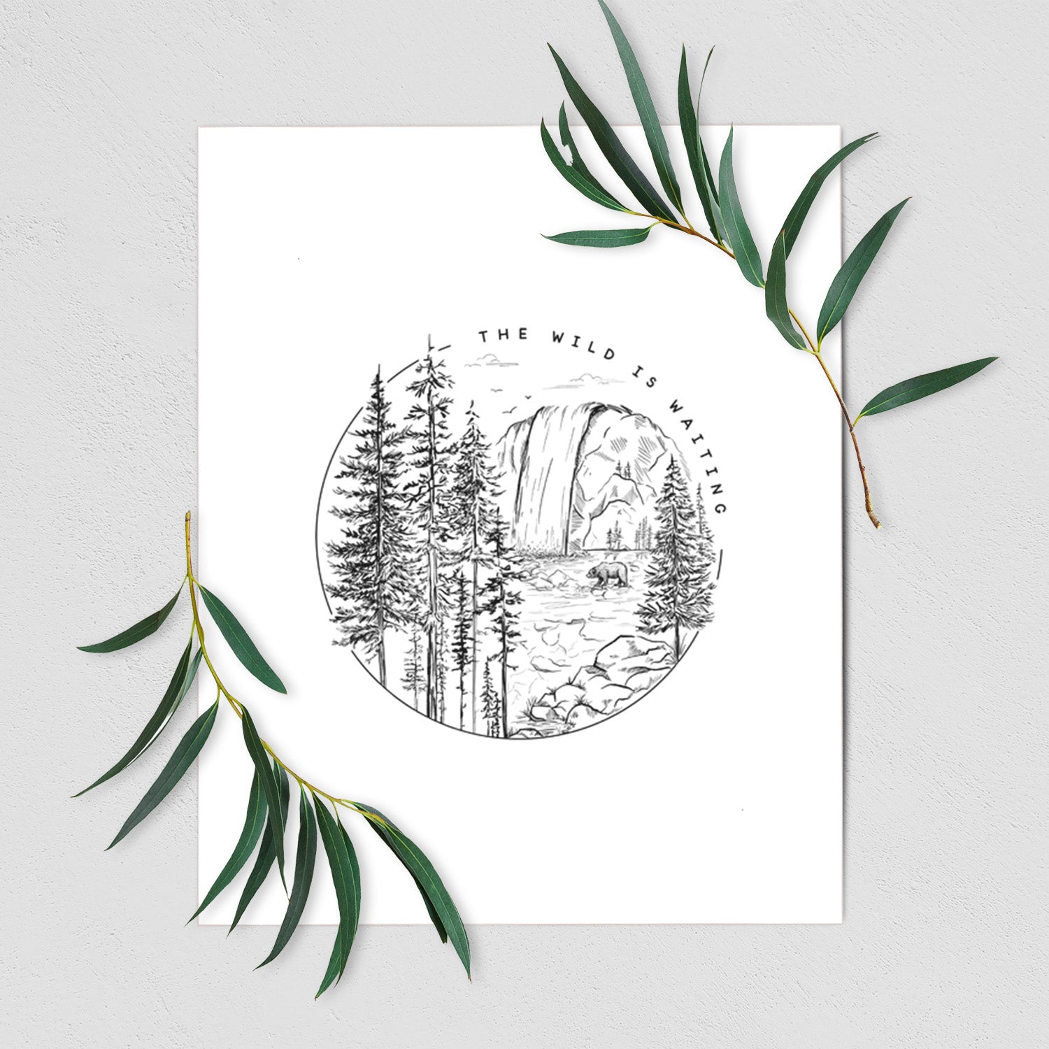 Art print of a round black ink scene of a waterfall, pine trees and a river, with the words The Wild Is Waiting, shown with eucalyptus leaves.
