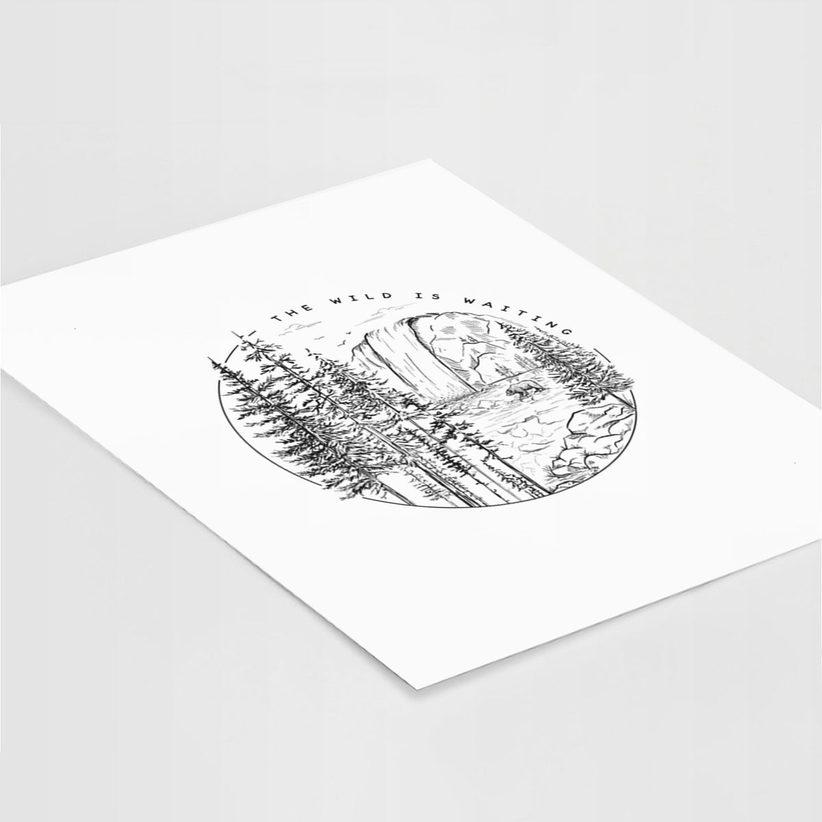 Art print of a round black ink scene of a waterfall, pine trees and a river, with the words The Wild Is Waiting, lying at an angle on a white surface.