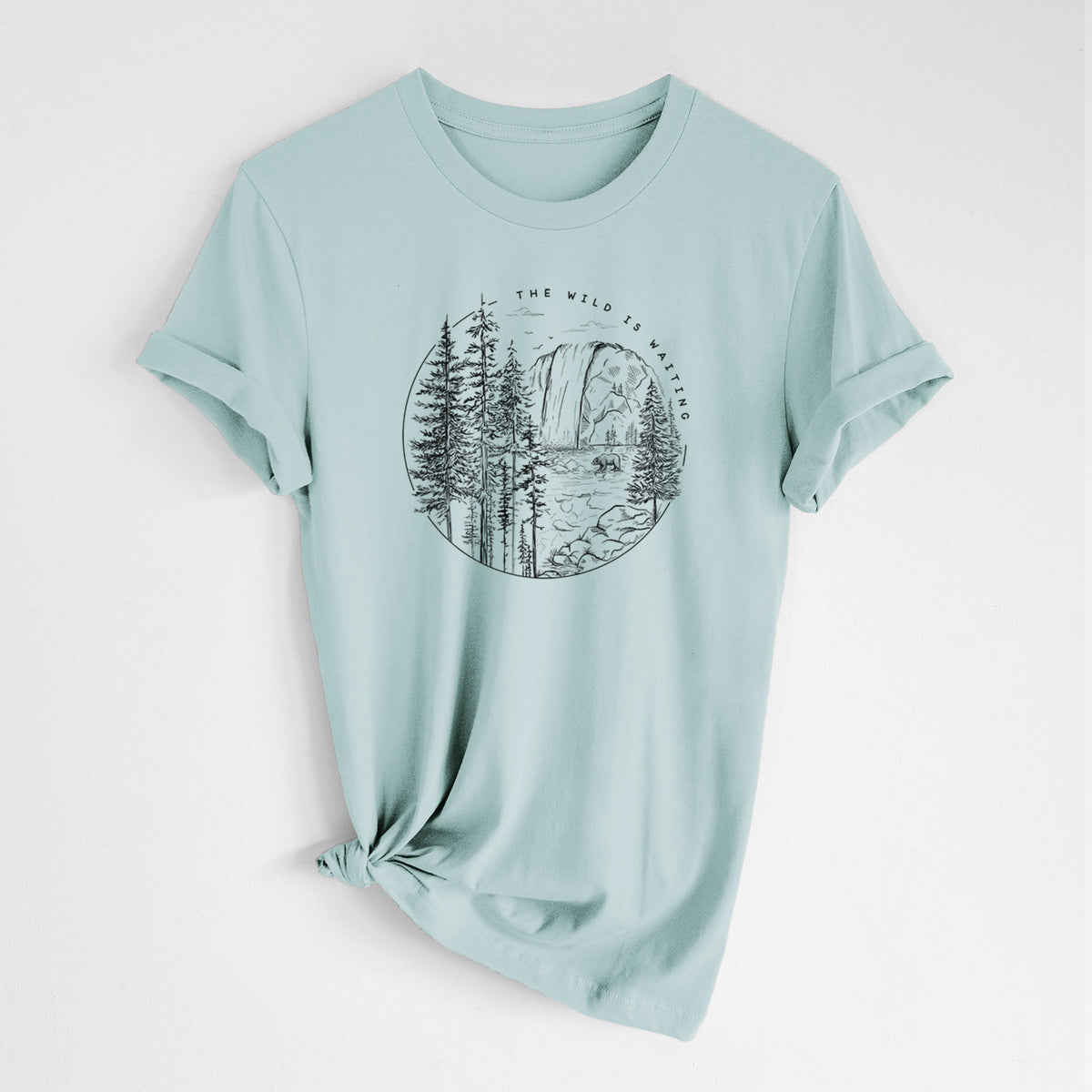 Dusty blue lightweight cotton t-shirt laid flat with the sleeves rolled, printed with a round black ink scene of a waterfall, pine trees and a river, with the words The Wild Is Waiting.