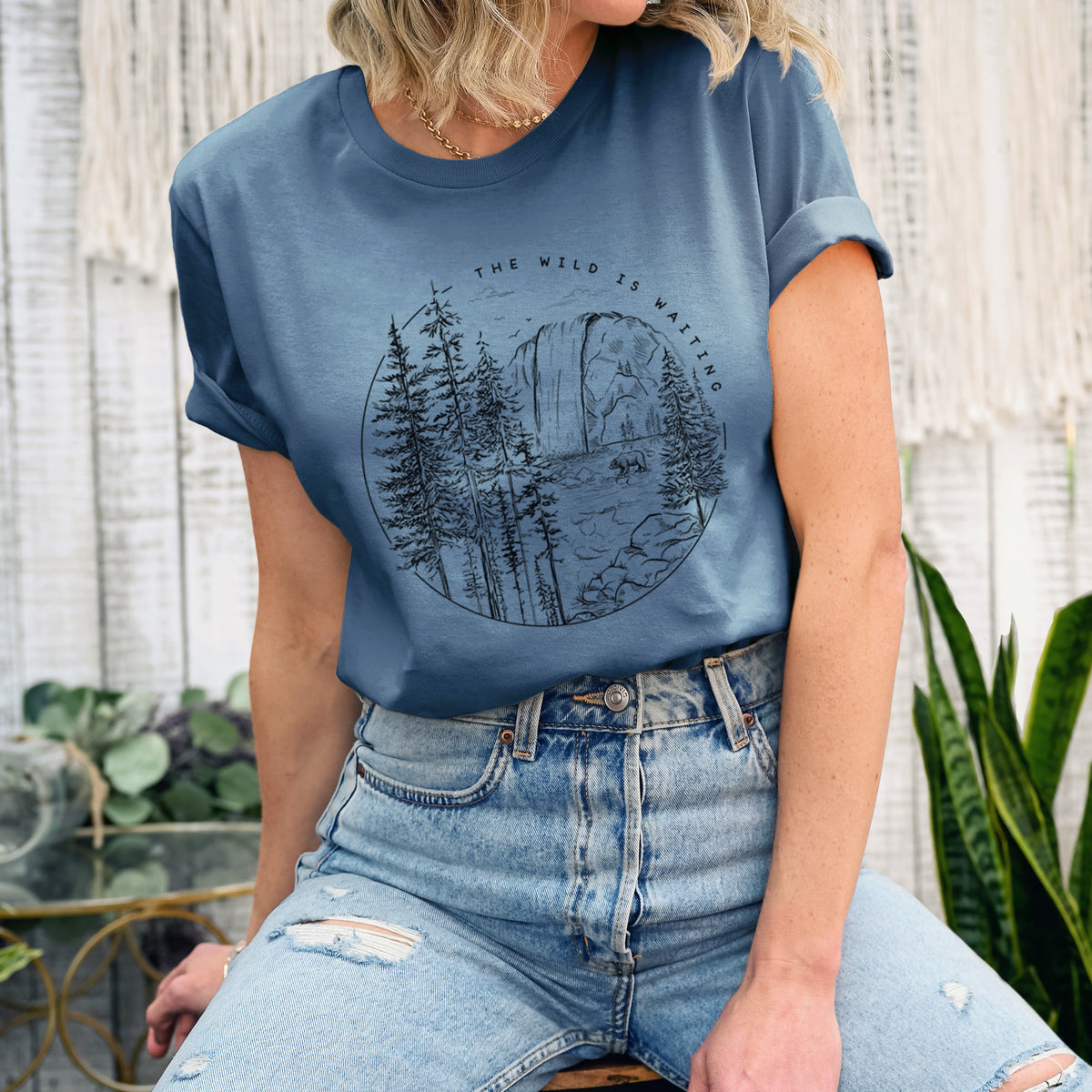 Woman wearing a steel blue lightweight cotton t-shirt printed with a round black ink scene of a waterfall, pine trees and a river, with the words The Wild Is Waiting.