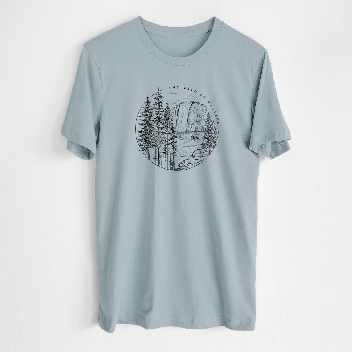Vintage denim lightweight cotton t-shirt laid flat, printed with a round black ink scene of a waterfall, pine trees and a river, with the words The Wild Is Waiting.