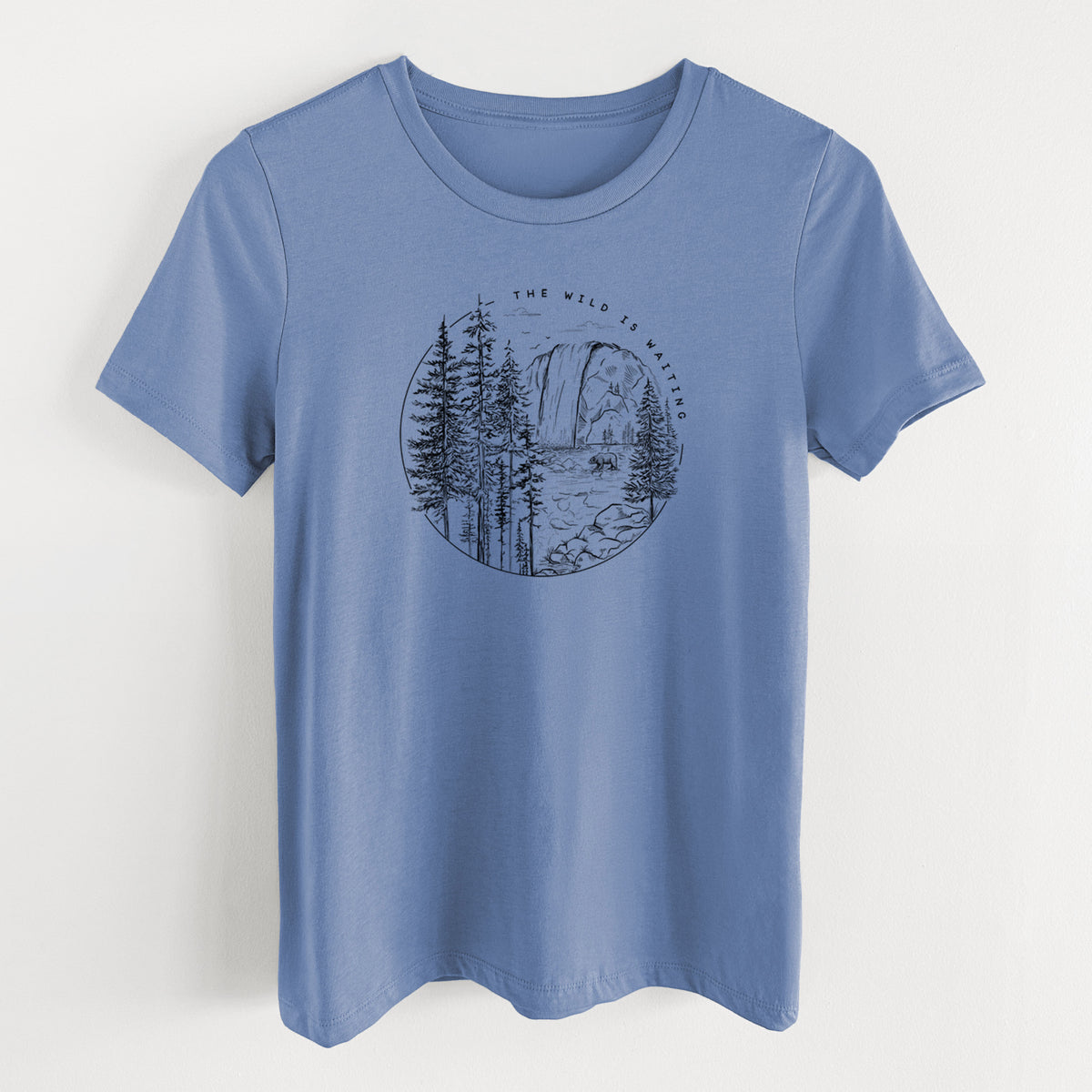 Blue bell women's relaxed fit cotton t-shirt laid flat, printed with a round black ink scene of a waterfall, pine trees and a river, with the words The Wild Is Waiting.