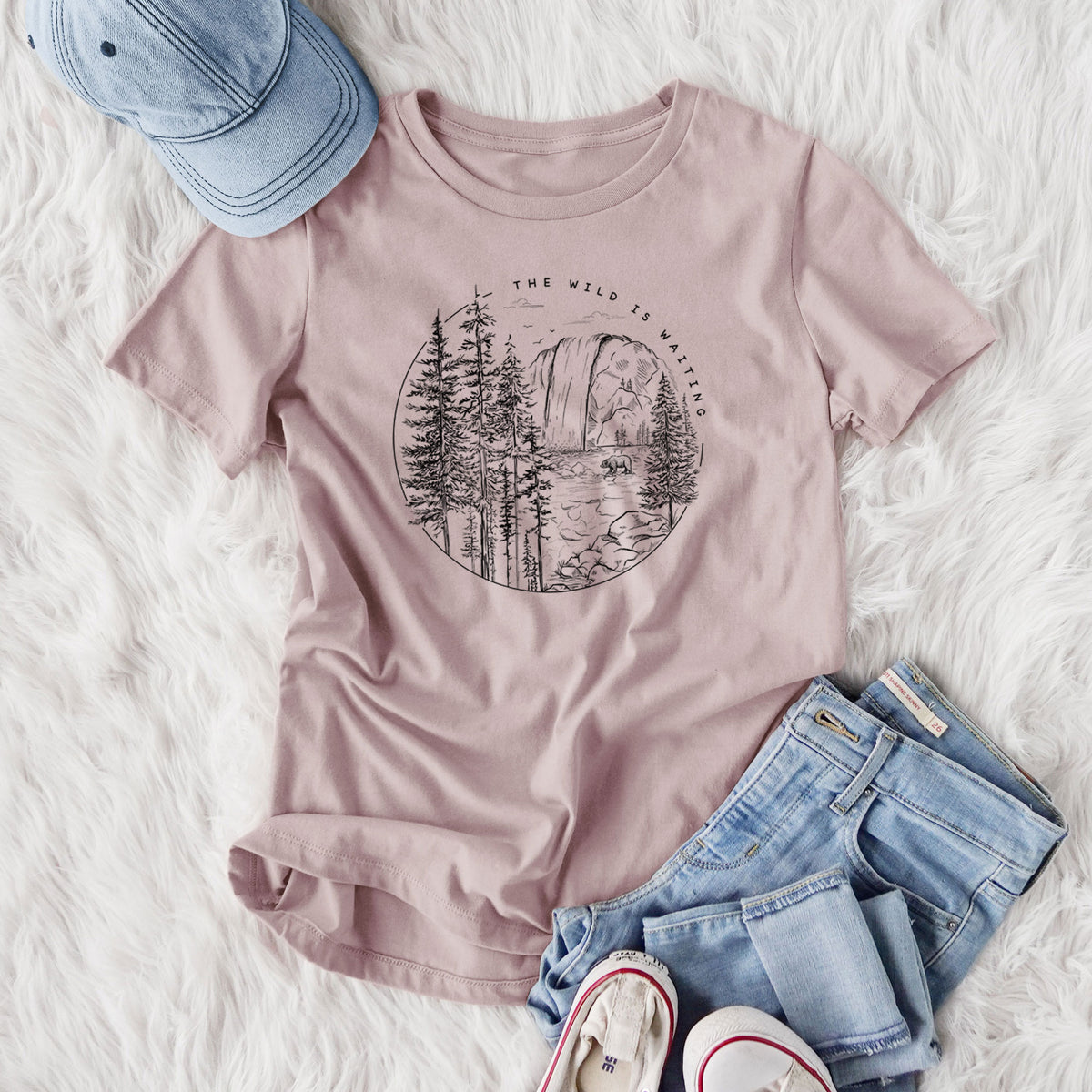 Women's relaxed fit cotton t-shirt printed with a round black ink scene of a waterfall, pine trees and a river, with the words The Wild Is Waiting, styled flat on a white faux fur rug with jeans.