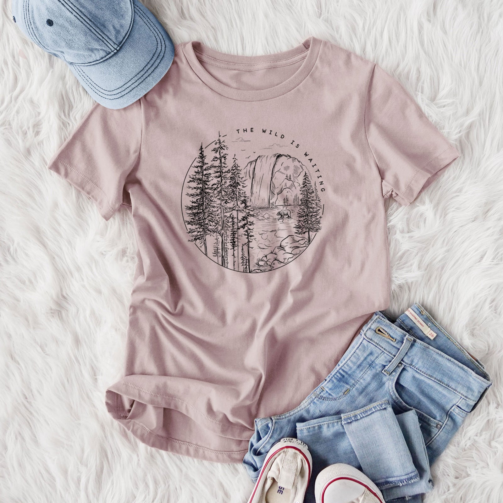 Women's relaxed fit cotton t-shirt printed with a round black ink scene of a waterfall, pine trees and a river, with the words The Wild Is Waiting, styled flat on a white faux fur rug with jeans.