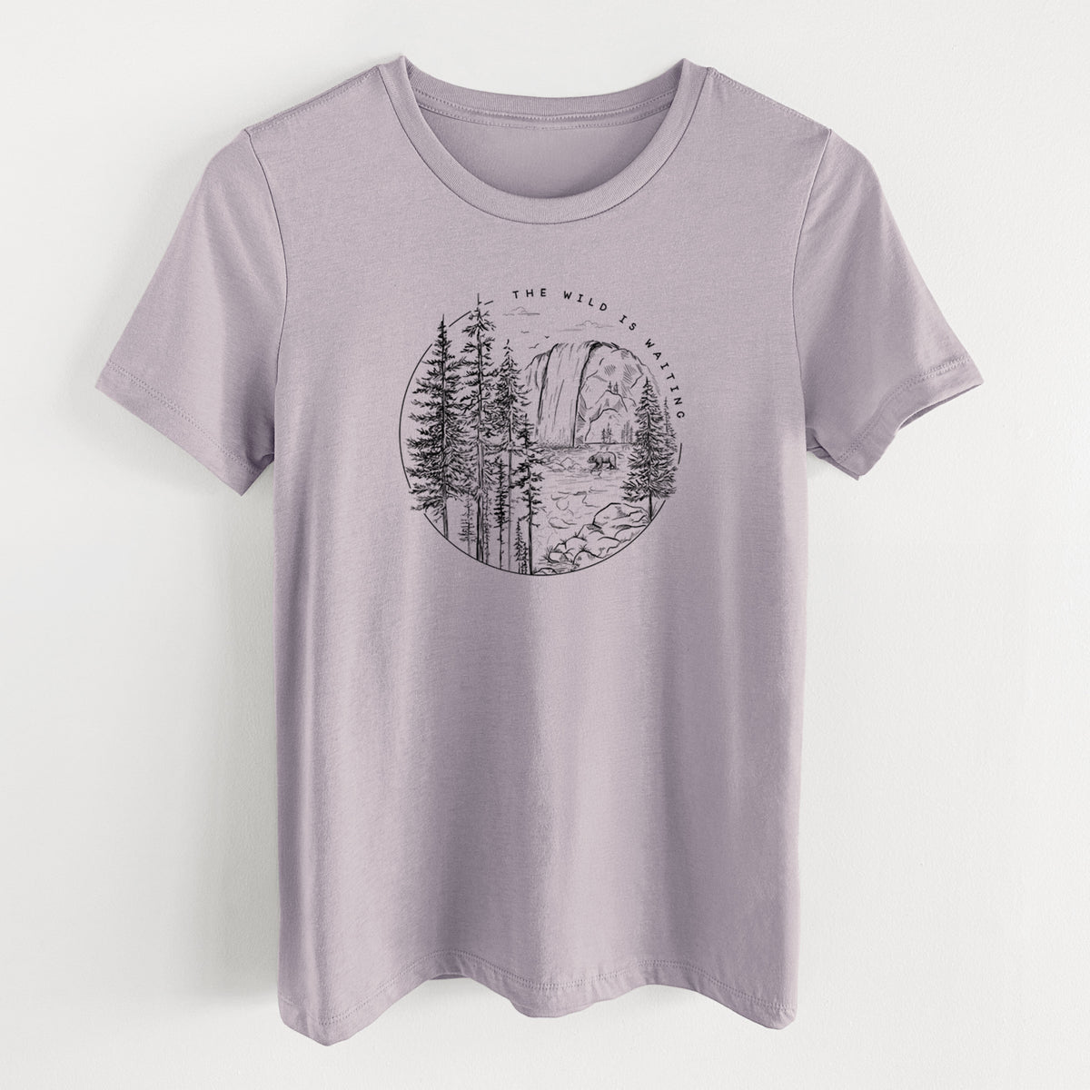 Light violet women's relaxed fit cotton t-shirt laid flat, printed with a round black ink scene of a waterfall, pine trees and a river, with the words The Wild Is Waiting.