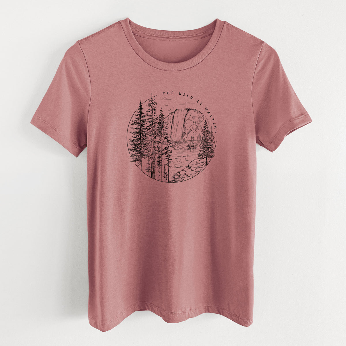 Mauve pink women's relaxed fit cotton t-shirt laid flat, printed with a round black ink scene of a waterfall, pine trees and a river, with the words The Wild Is Waiting.