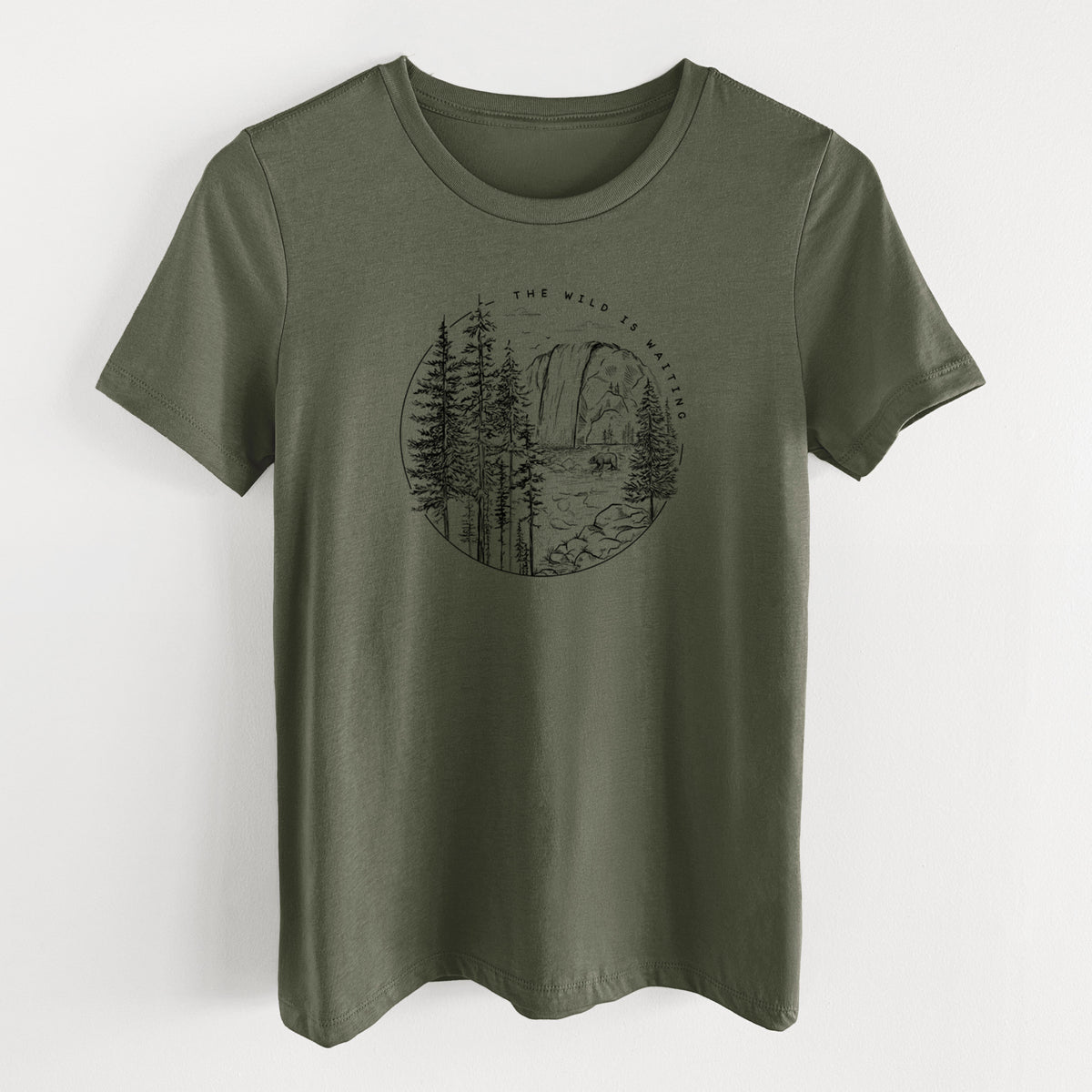 Military green women's relaxed fit cotton t-shirt laid flat, printed with a round black ink scene of a waterfall, pine trees and a river, with the words The Wild Is Waiting.