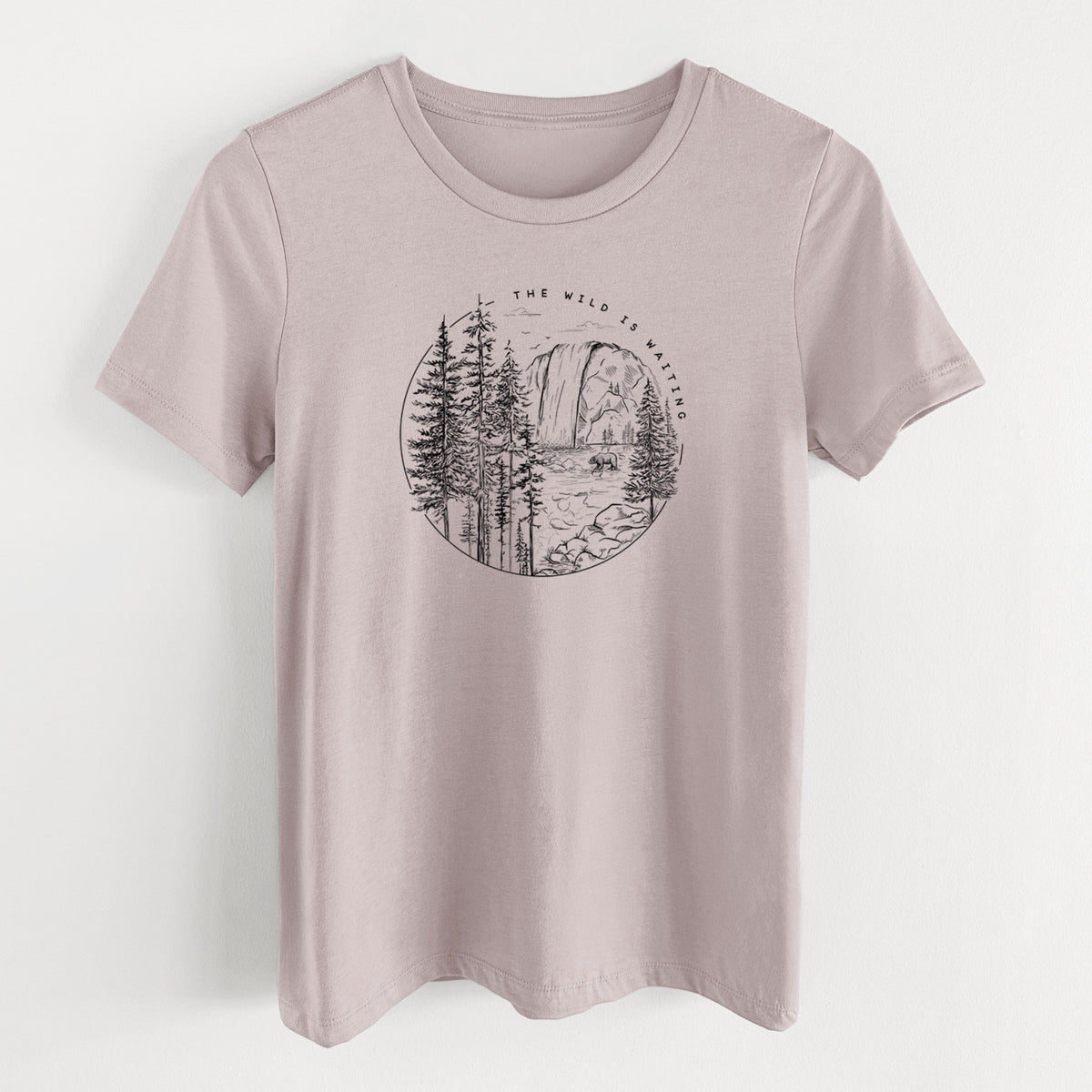 Pink gravel women's relaxed fit cotton t-shirt laid flat, printed with a round black ink scene of a waterfall, pine trees and a river, with the words The Wild Is Waiting.