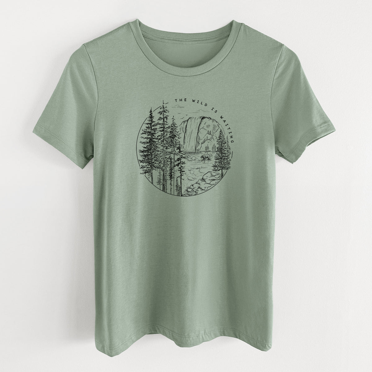 Sage green women's relaxed fit cotton t-shirt laid flat, printed with a round black ink scene of a waterfall, pine trees and a river, with the words The Wild Is Waiting.
