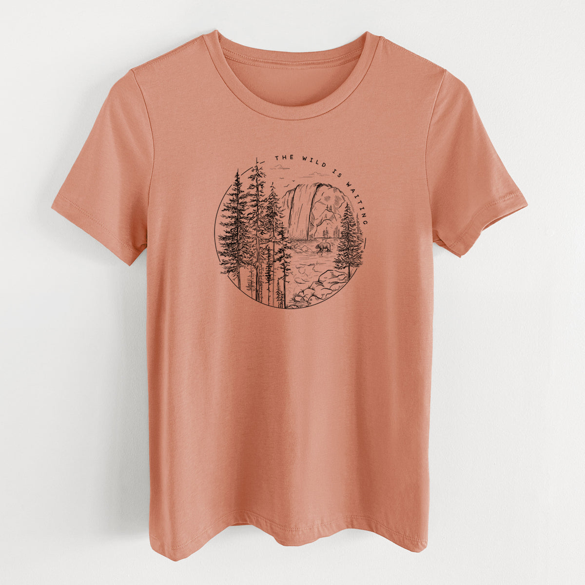 Terracotta orange women's relaxed fit cotton t-shirt laid flat, printed with a round black ink scene of a waterfall, pine trees and a river, with the words The Wild Is Waiting.