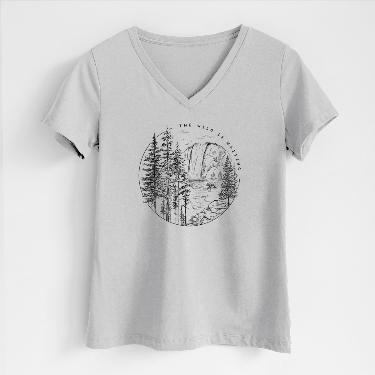 Lunar grey women's relaxed fit cotton V-neck t-shirt laid flat, printed with a round black ink scene of a waterfall, pine trees and a river, with the words The Wild Is Waiting.