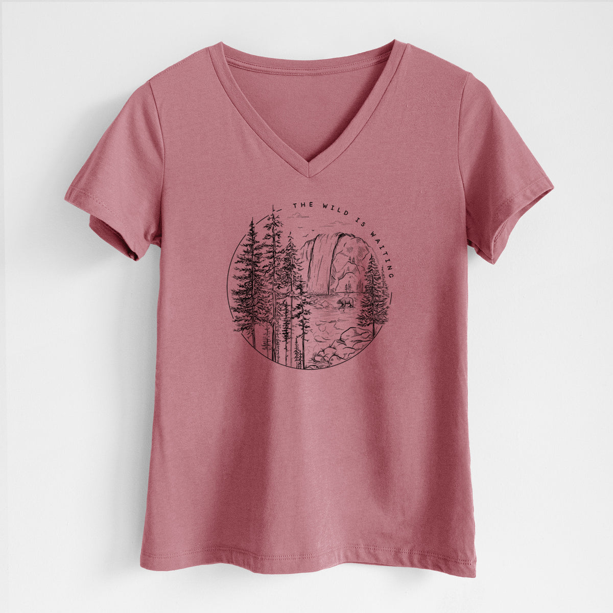 Mauve pink women's relaxed fit cotton V-neck t-shirt laid flat, printed with a round black ink scene of a waterfall, pine trees and a river, with the words The Wild Is Waiting.