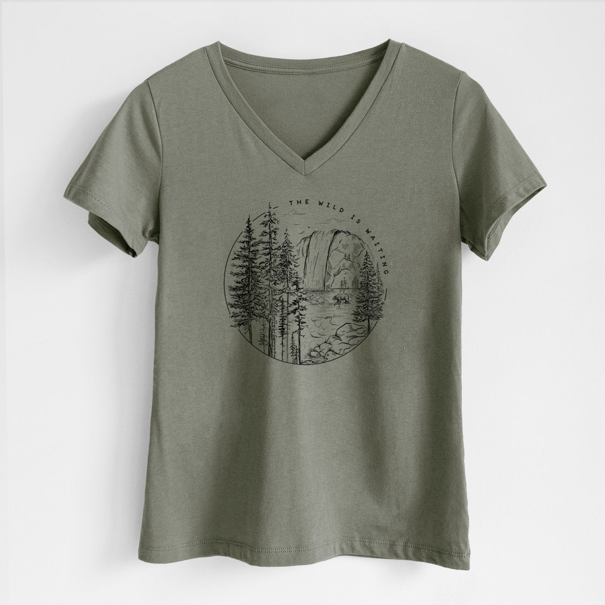 Military green women's relaxed fit cotton V-neck t-shirt laid flat, printed with a round black ink scene of a waterfall, pine trees and a river, with the words The Wild Is Waiting.