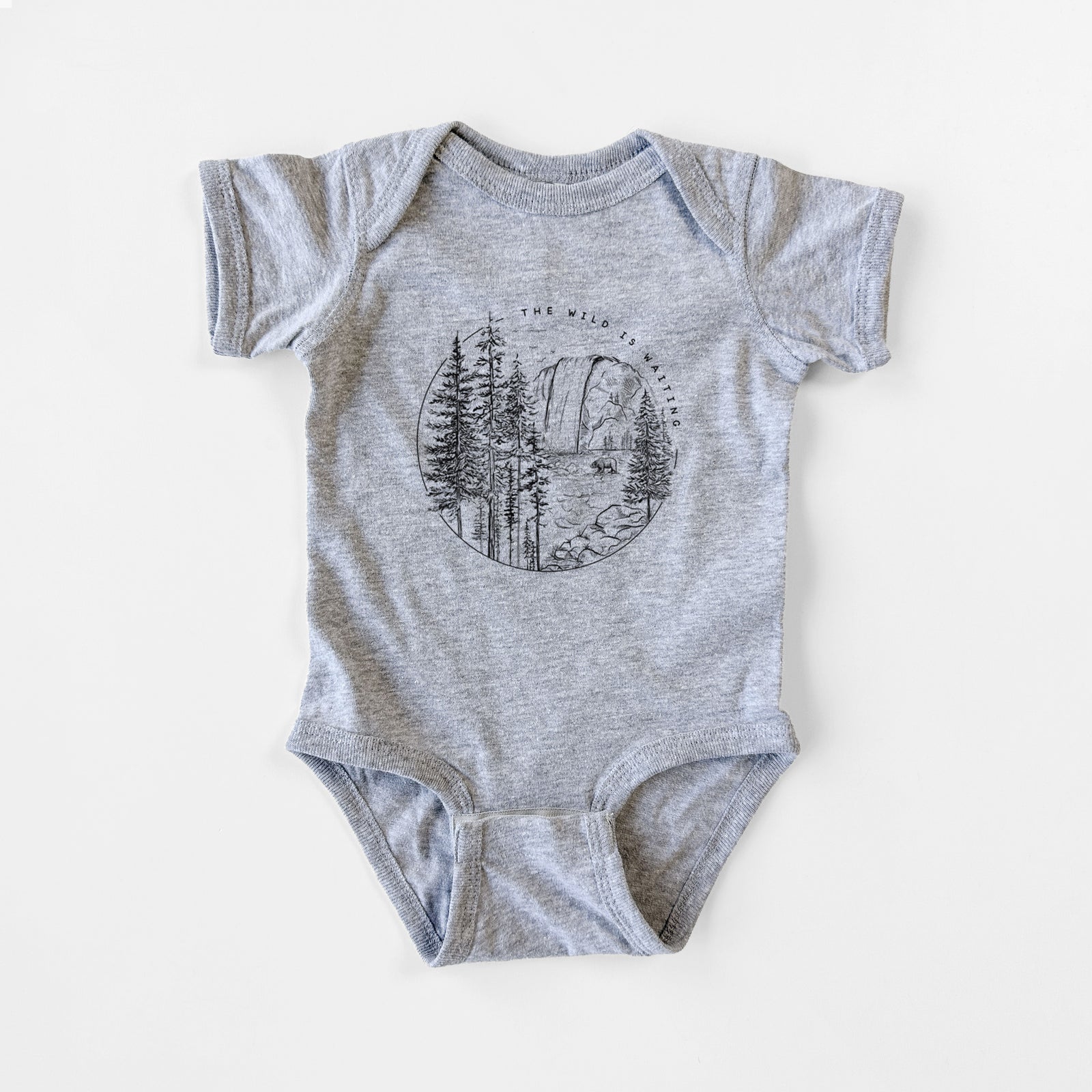 Grey baby bodysuit laid flat, printed with a round black ink scene of a waterfall, pine trees and a river, with the words The Wild Is Waiting.
