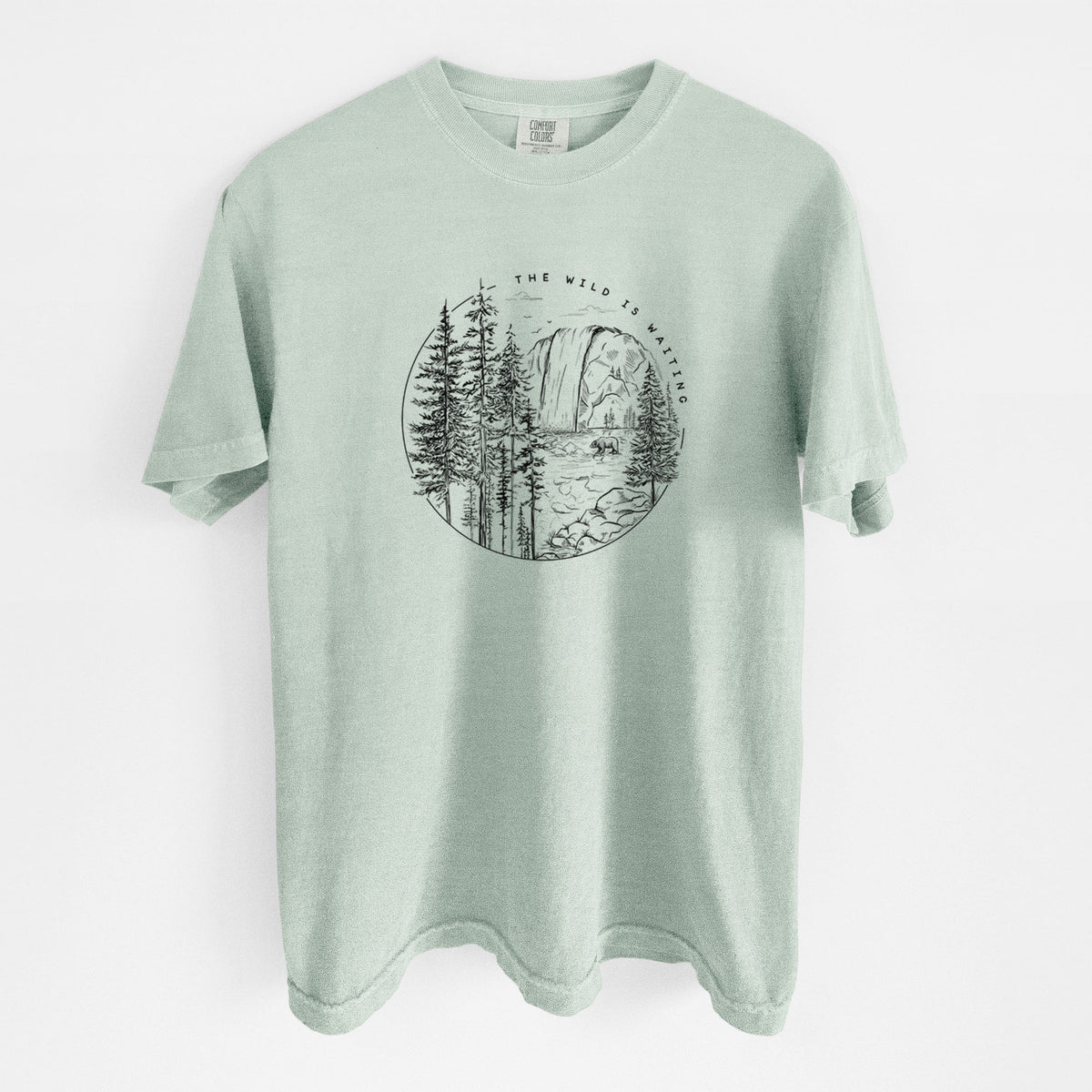 Bay green Comfort Colors t-shirt laid flat, printed with a round black ink scene of a waterfall, pine trees and a river, with the words The Wild Is Waiting.