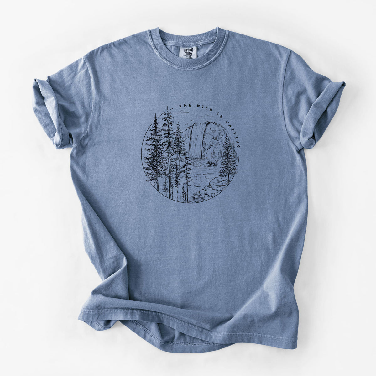 Blue jean Comfort Colors t-shirt laid flat, printed with a round black ink scene of a waterfall, pine trees and a river, with the words The Wild Is Waiting.
