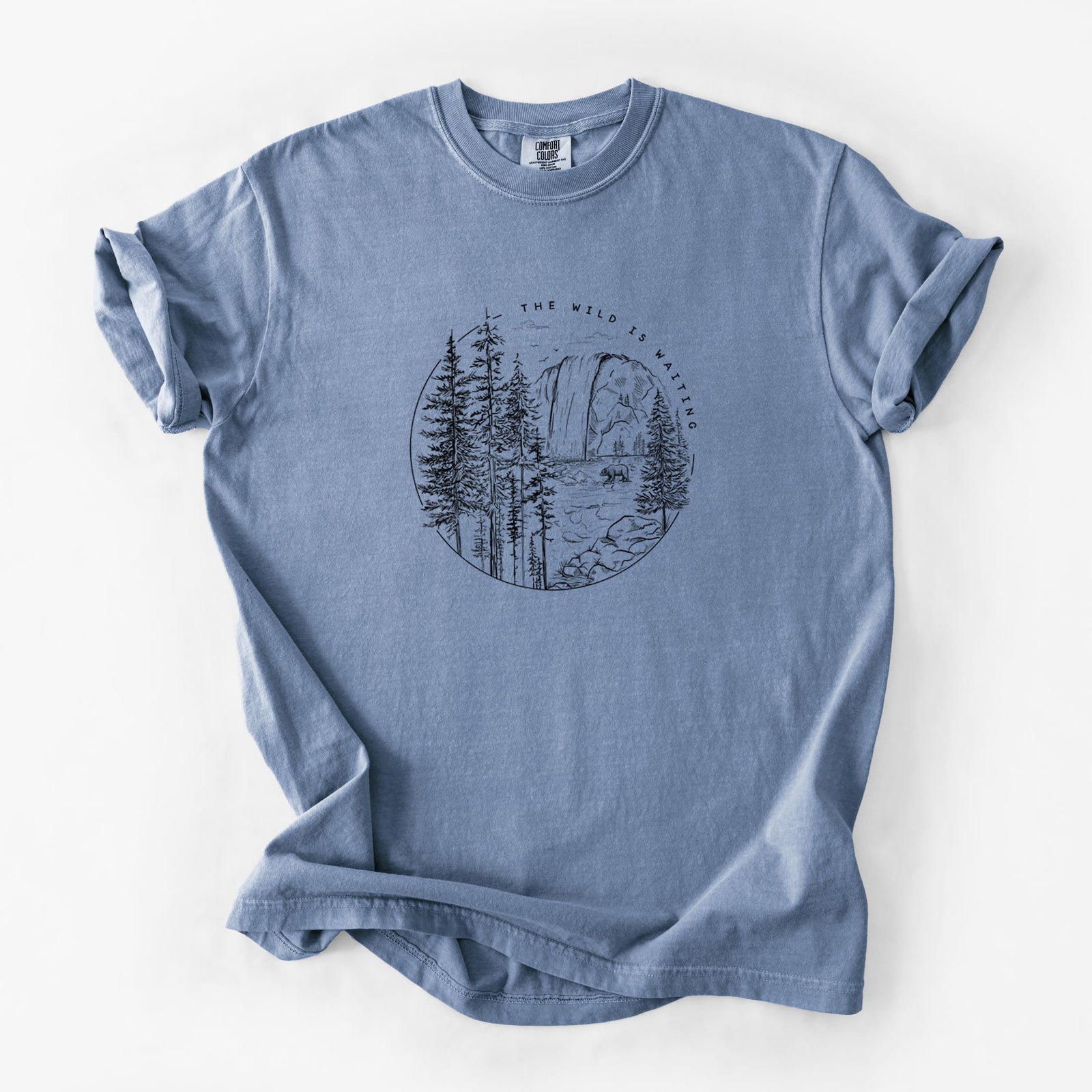 Blue jean Comfort Colors t-shirt laid flat, printed with a round black ink scene of a waterfall, pine trees and a river, with the words The Wild Is Waiting.