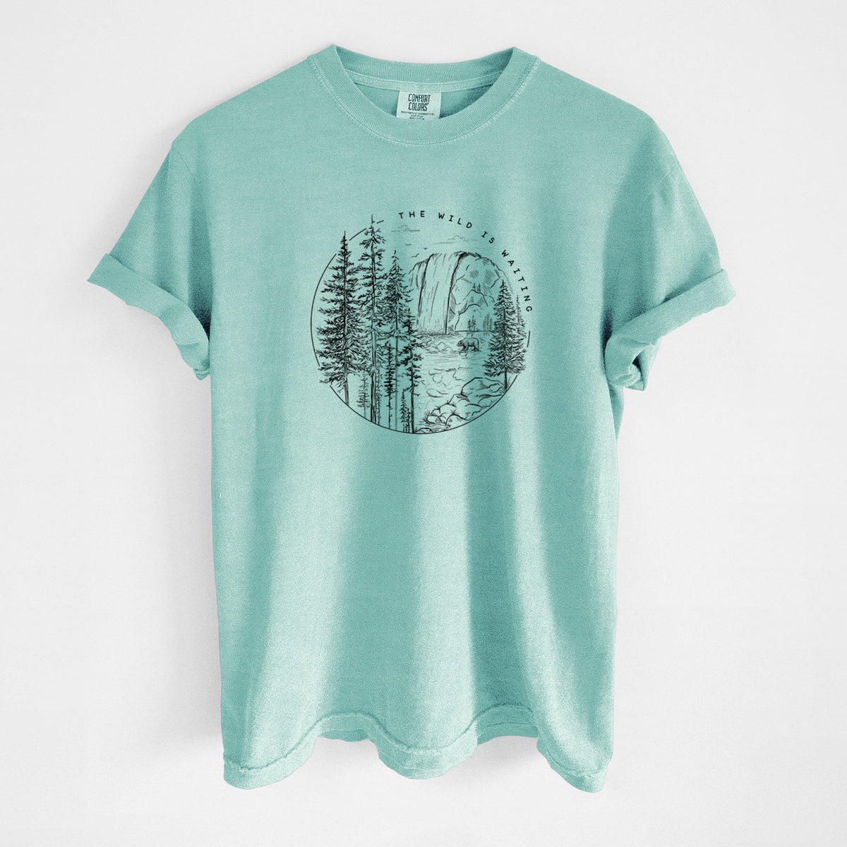 Chalky mint Comfort Colors t-shirt laid flat, printed with a round black ink scene of a waterfall, pine trees and a river, with the words The Wild Is Waiting.