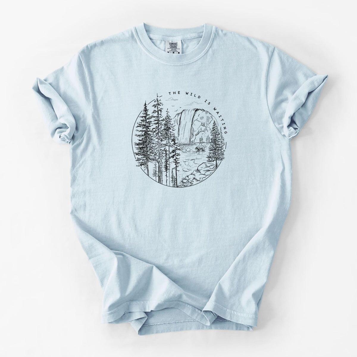 Chambray blue Comfort Colors t-shirt laid flat, printed with a round black ink scene of a waterfall, pine trees and a river, with the words The Wild Is Waiting.
