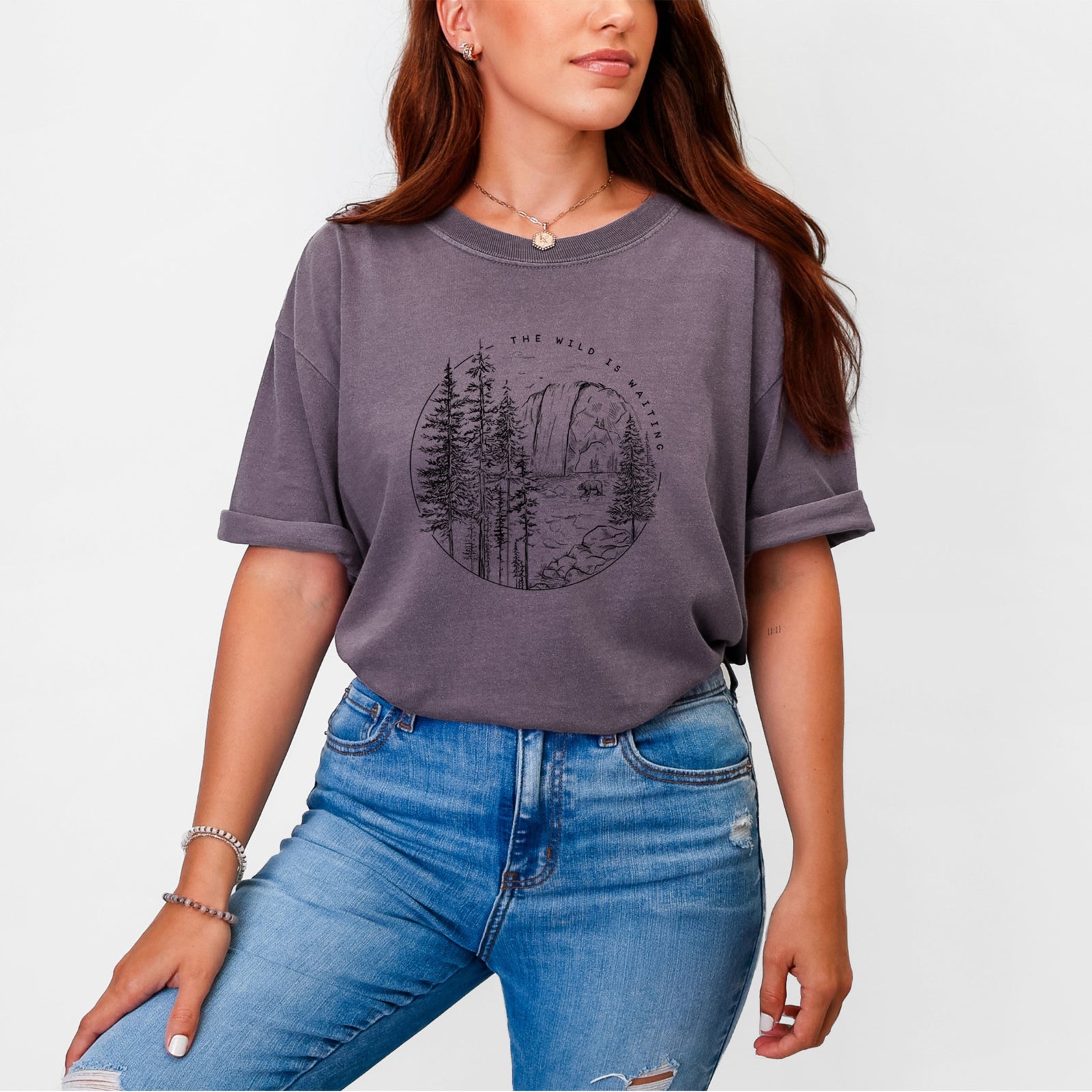 Blue jean Comfort Colors t-shirt laid flat, printed with a round black ink scene of a waterfall, pine trees and a river, with the words The Wild Is Waiting.