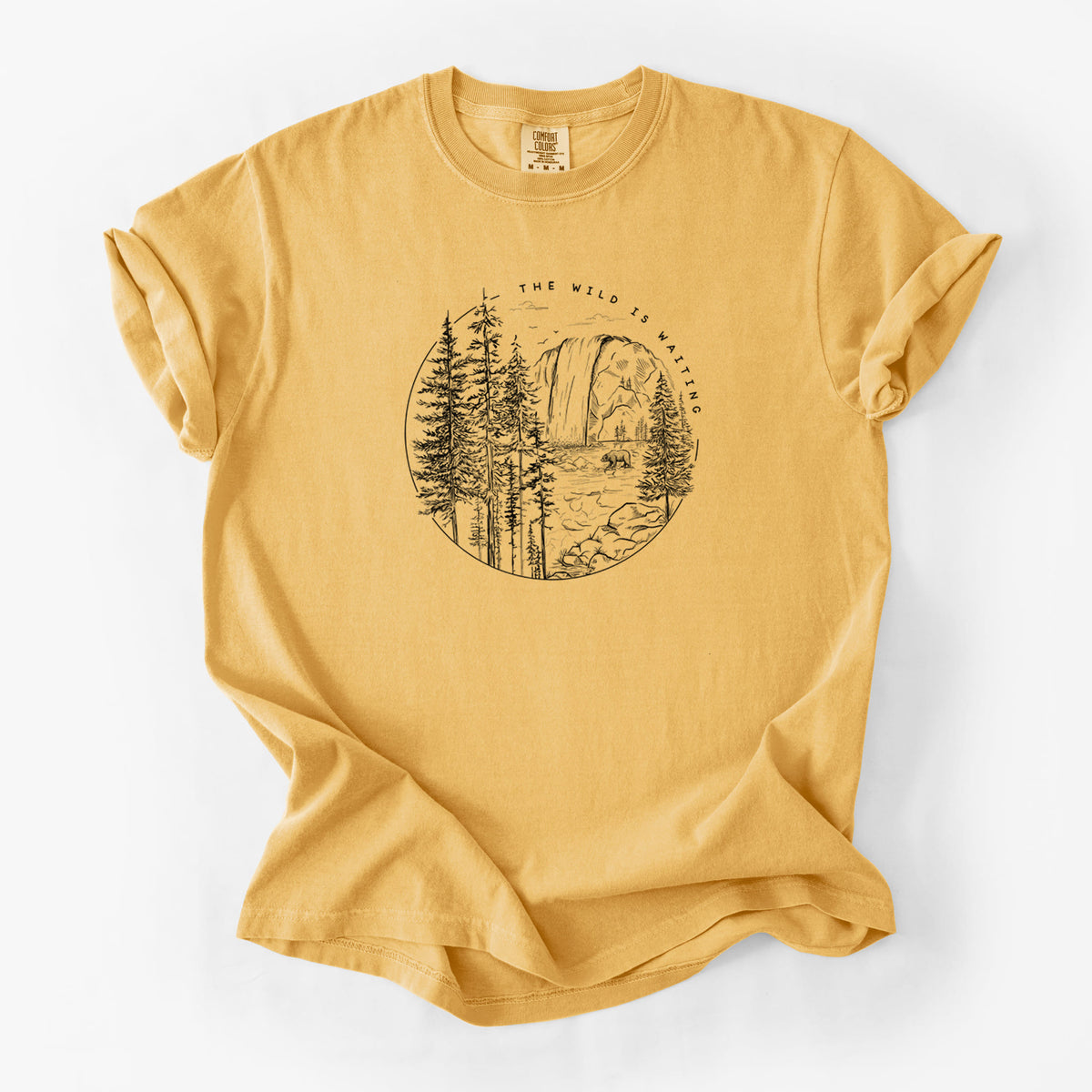 Golden wheat yellow Comfort Colors t-shirt laid flat, printed with a round black ink scene of a waterfall, pine trees and a river, with the words The Wild Is Waiting.