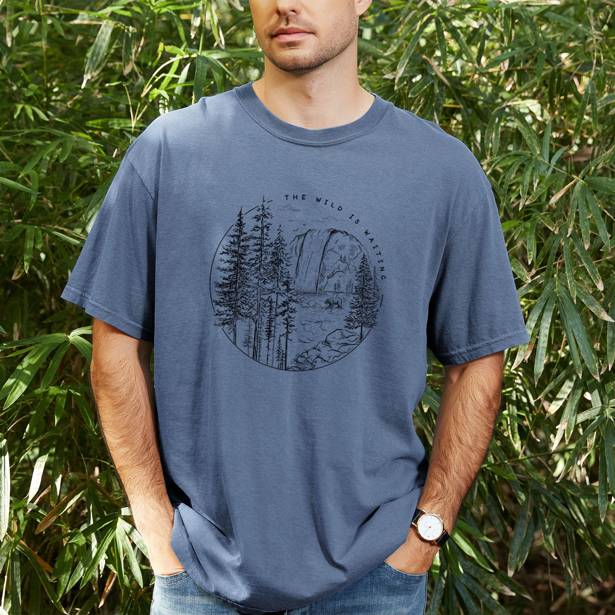 Man wearing a Comfort Colors t-shirt printed with a round black ink scene of a waterfall, pine trees and a river, with the words The Wild Is Waiting, outdoors.