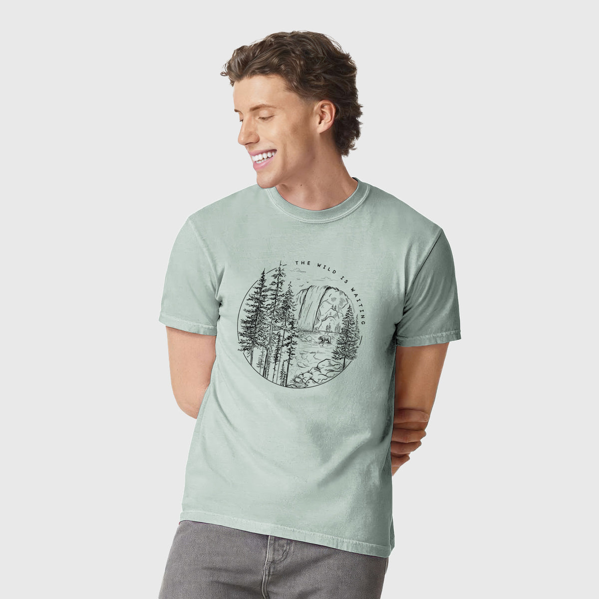 Man wearing a Comfort Colors t-shirt printed with a round black ink scene of a waterfall, pine trees and a river, with the words The Wild Is Waiting.
