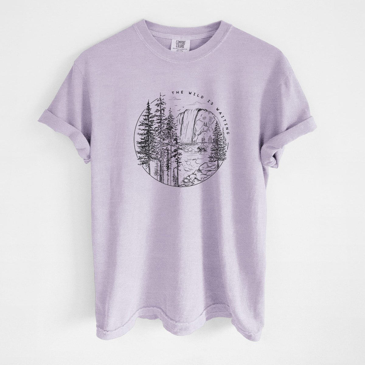 Orchid purple Comfort Colors t-shirt laid flat, printed with a round black ink scene of a waterfall, pine trees and a river, with the words The Wild Is Waiting.
