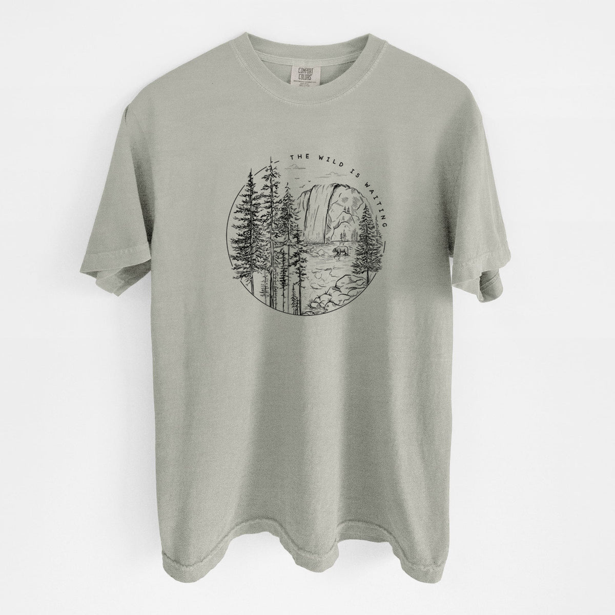 Sandstone beige Comfort Colors t-shirt laid flat, printed with a round black ink scene of a waterfall, pine trees and a river, with the words The Wild Is Waiting.