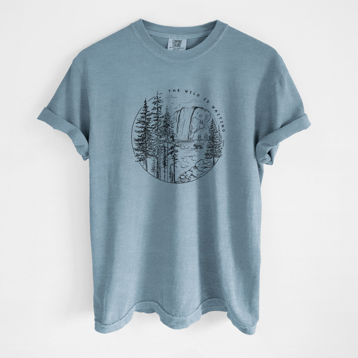 Steel blue Comfort Colors t-shirt laid flat, printed with a round black ink scene of a waterfall, pine trees and a river, with the words The Wild Is Waiting.