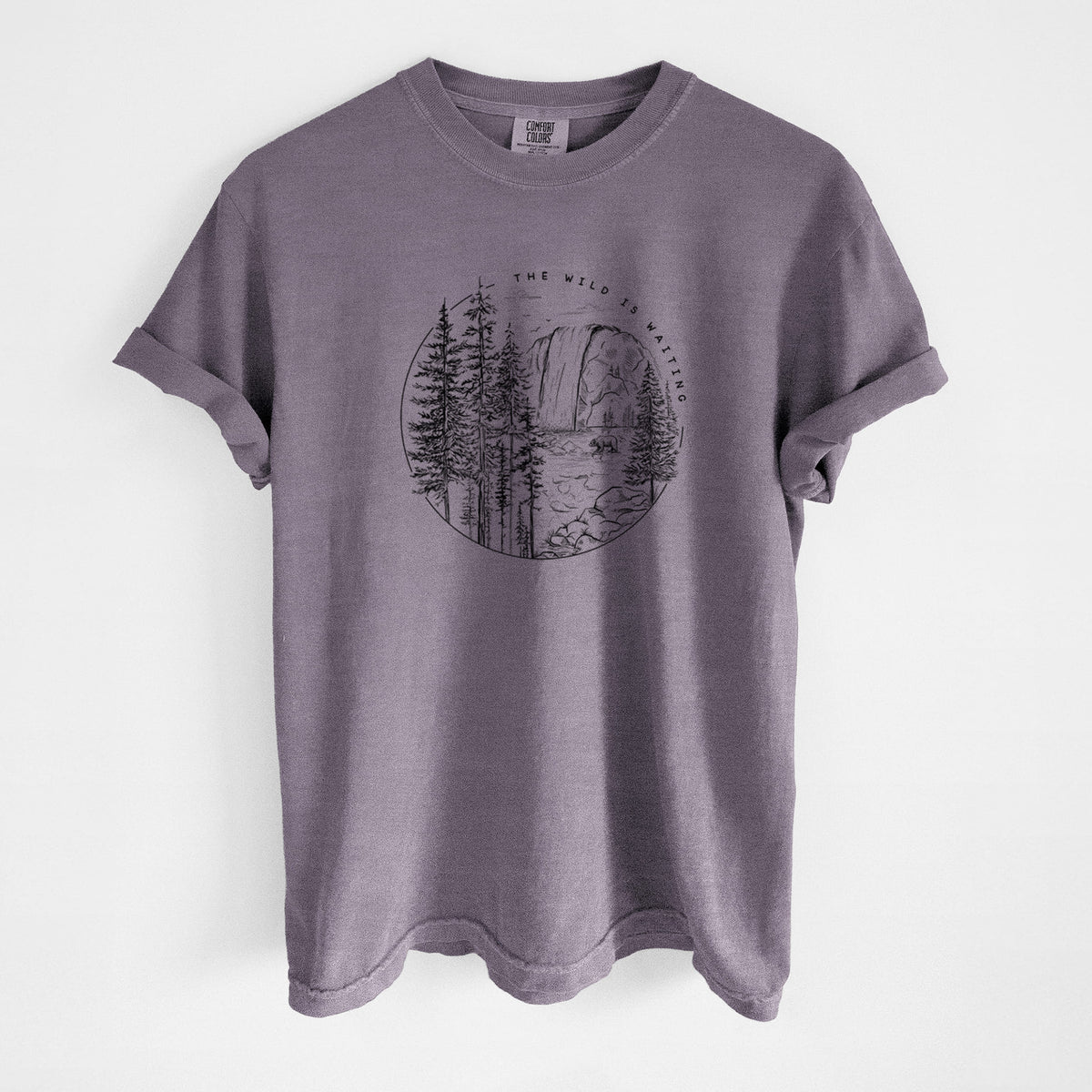Wine red Comfort Colors t-shirt laid flat, printed with a round black ink scene of a waterfall, pine trees and a river, with the words The Wild Is Waiting.