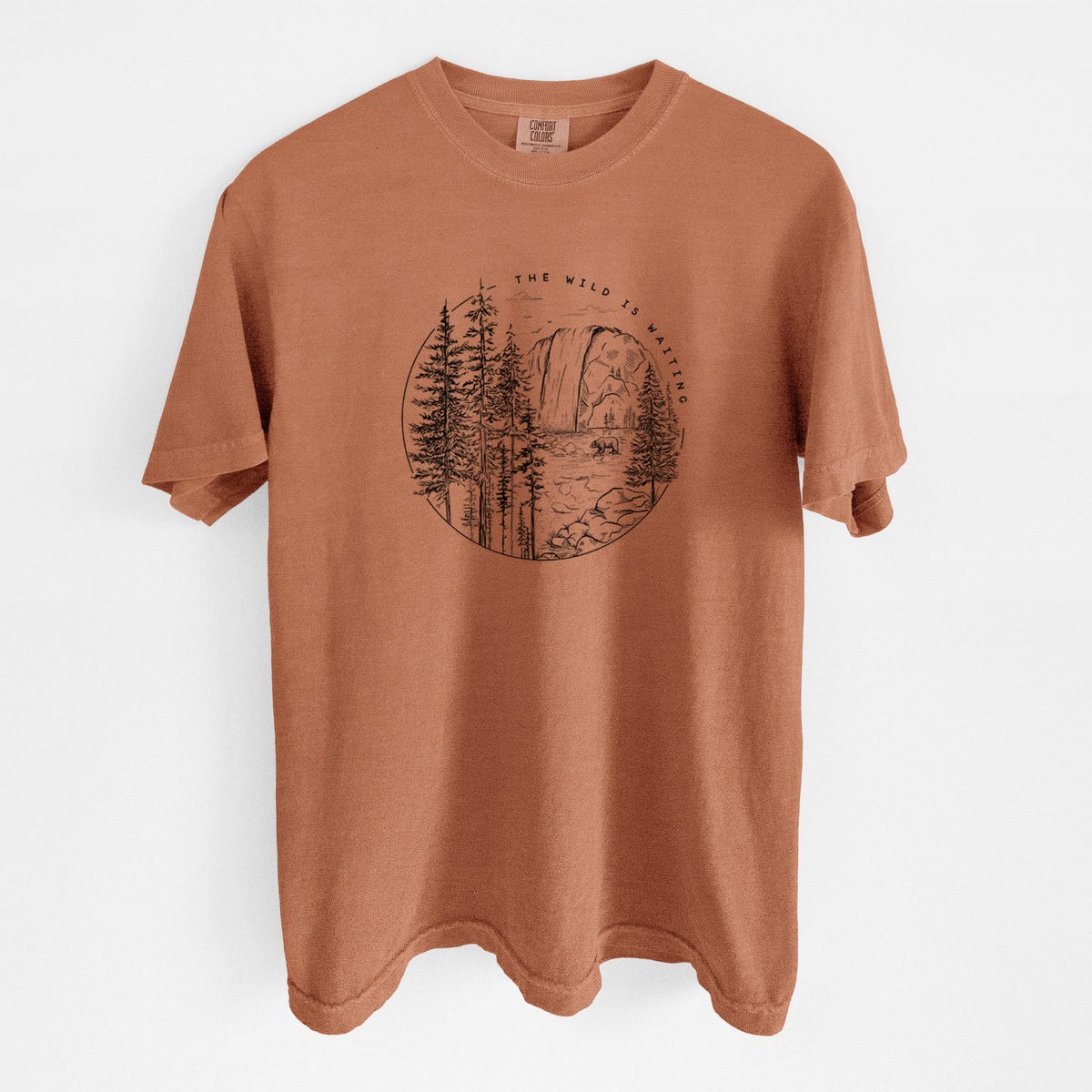 Yam burnt orange Comfort Colors t-shirt laid flat, printed with a round black ink scene of a waterfall, pine trees and a river, with the words The Wild Is Waiting.