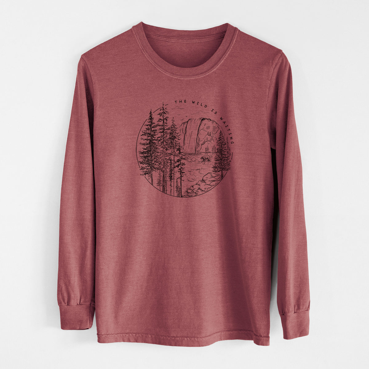 Brick red Comfort Colors long sleeve shirt laid flat, printed with a round black ink scene of a waterfall, pine trees and a river, with the words The Wild Is Waiting.