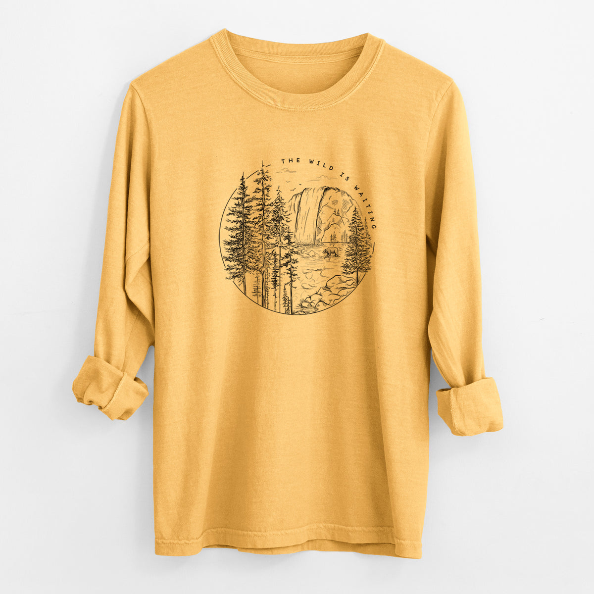 Golden wheat yellow Comfort Colors long sleeve shirt laid flat with the cuffs rolled, printed with a round black ink scene of a waterfall, pine trees and a river, with the words The Wild Is Waiting.