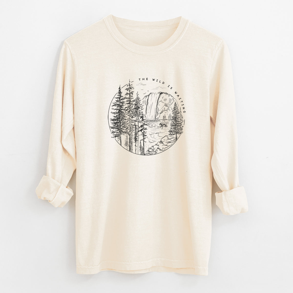 Ivory Comfort Colors long sleeve shirt laid flat with the cuffs rolled, printed with a round black ink scene of a waterfall, pine trees and a river, with the words The Wild Is Waiting.