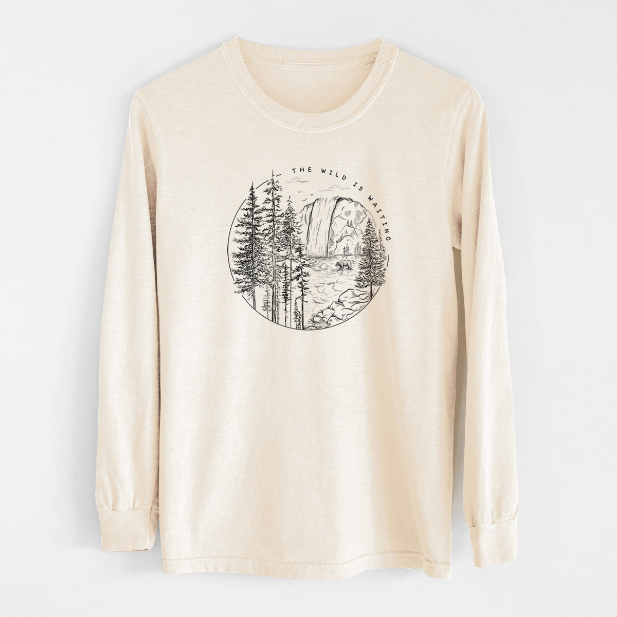 Ivory Comfort Colors long sleeve shirt laid flat, printed with a round black ink scene of a waterfall, pine trees and a river, with the words The Wild Is Waiting.
