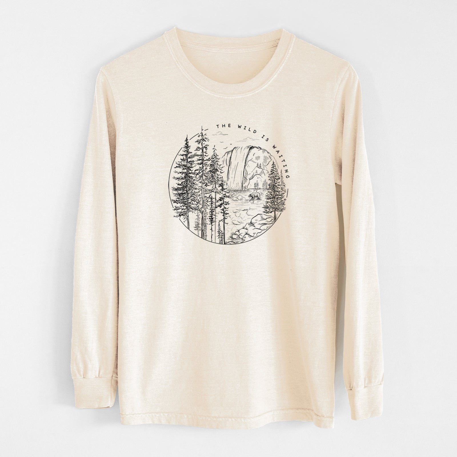 Ivory Comfort Colors long sleeve shirt laid flat, printed with a round black ink scene of a waterfall, pine trees and a river, with the words The Wild Is Waiting.