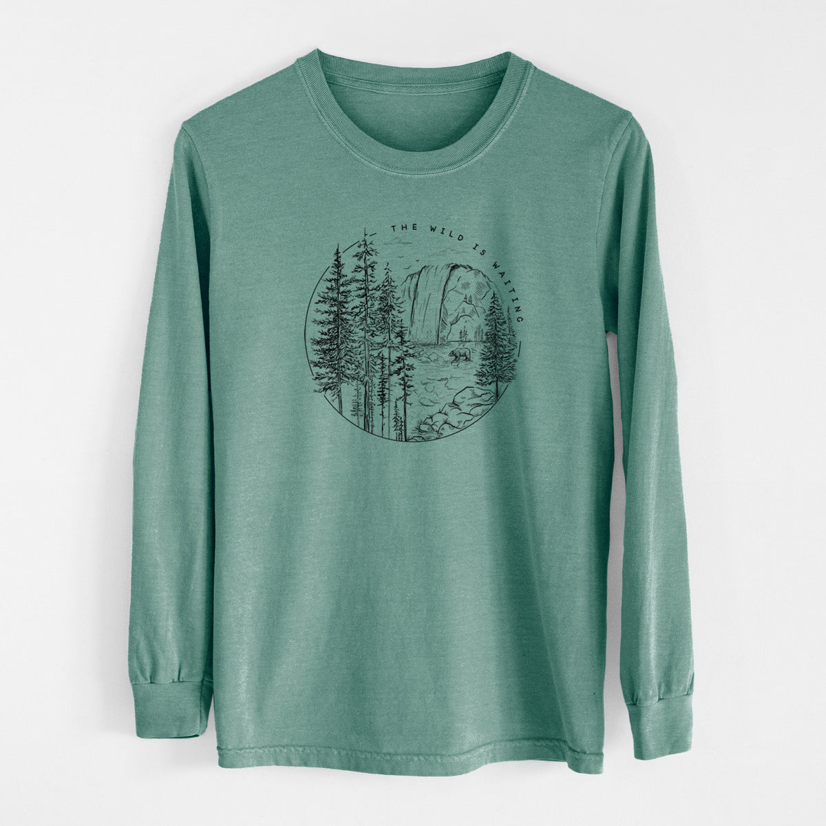 Pine green Comfort Colors long sleeve shirt laid flat, printed with a round black ink scene of a waterfall, pine trees and a river, with the words The Wild Is Waiting.