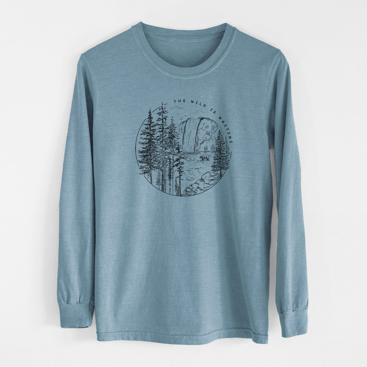 Steel blue Comfort Colors long sleeve shirt laid flat, printed with a round black ink scene of a waterfall, pine trees and a river, with the words The Wild Is Waiting.